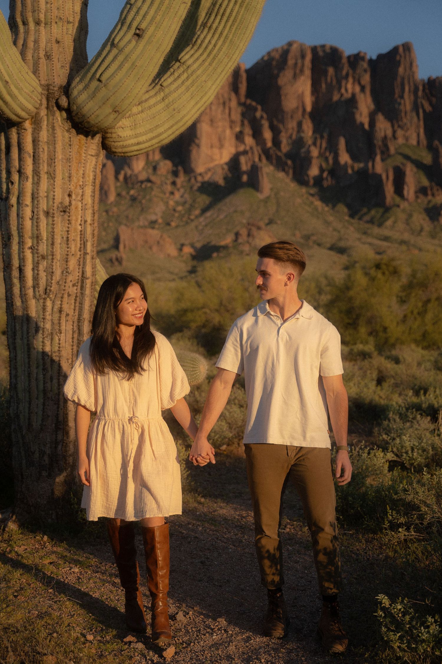 A couple holds hands while walking along a desert trail with a saguaro cactus and mountain backdrop at sunset.