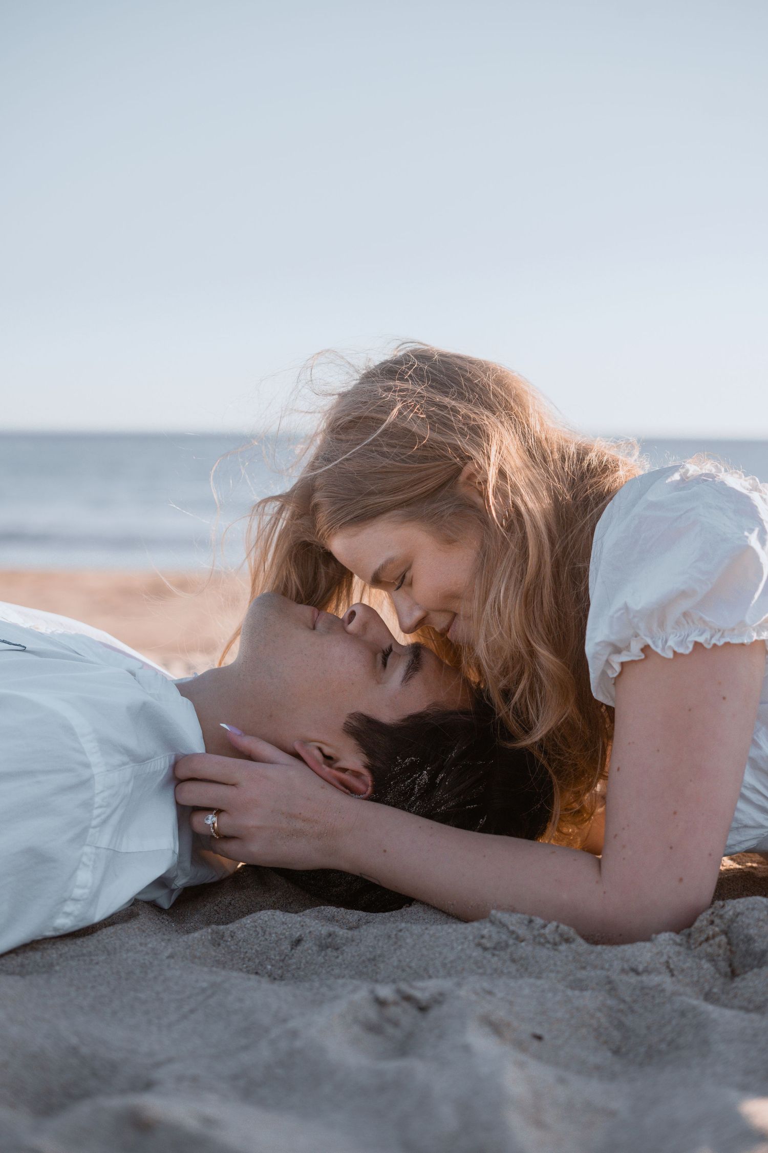 A romantic couple shares an intimate moment while laying on the beach sand at sunset.