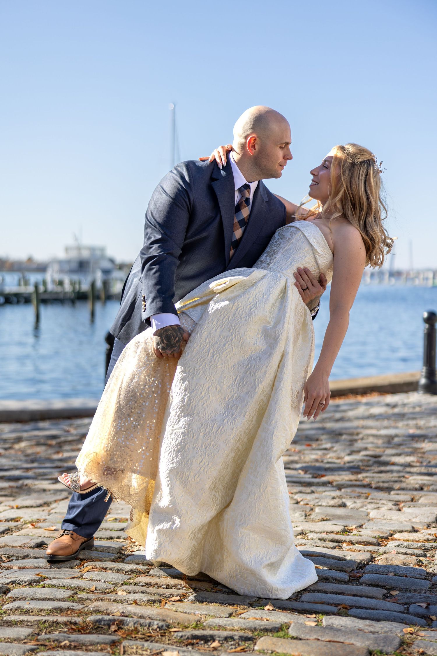Groom dips bride in white gown on cobblestone waterfront dock under blue sky.