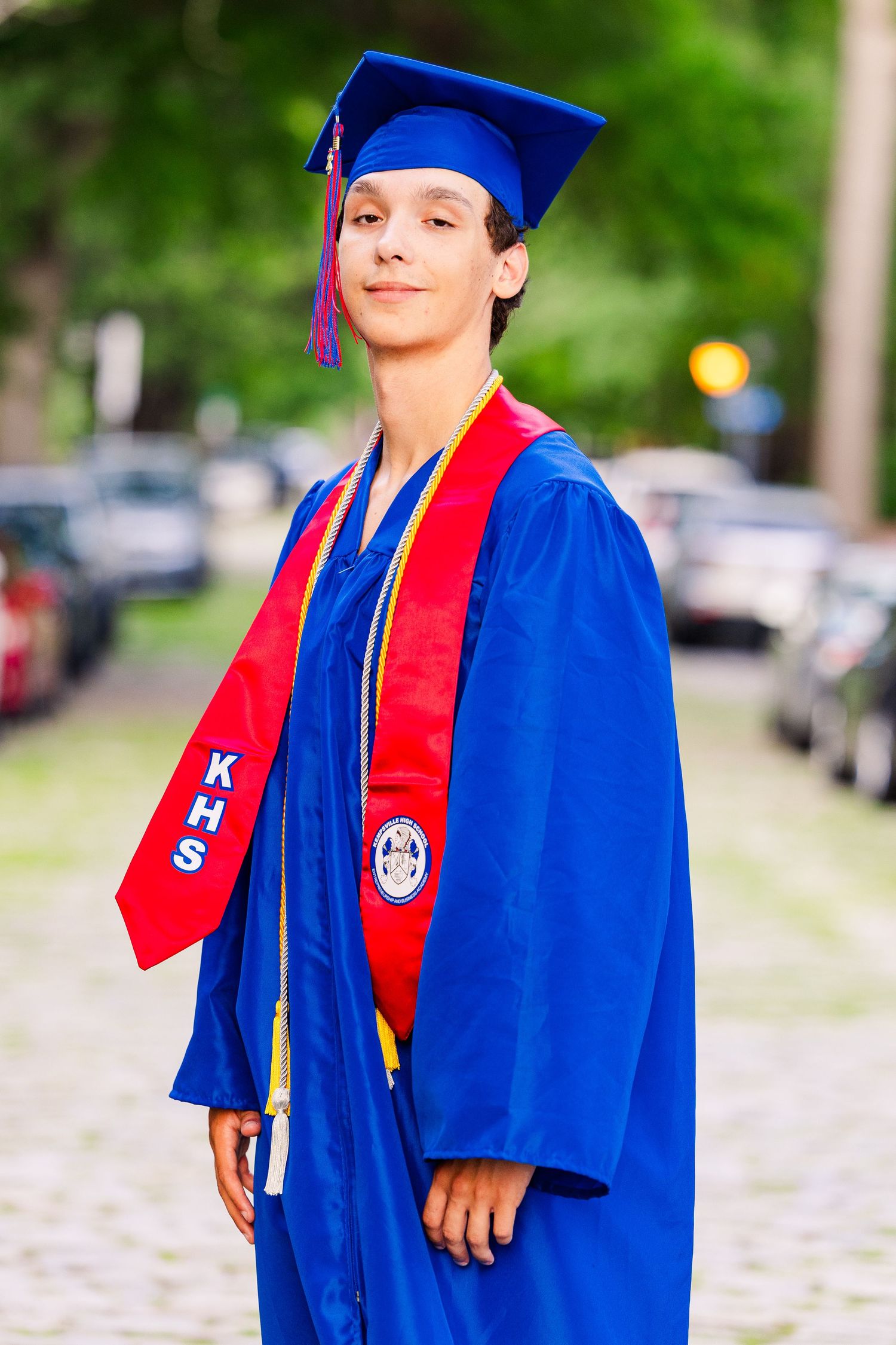 Young graduate in blue cap and gown with red KHS sash poses outdoors on cobblestone street.