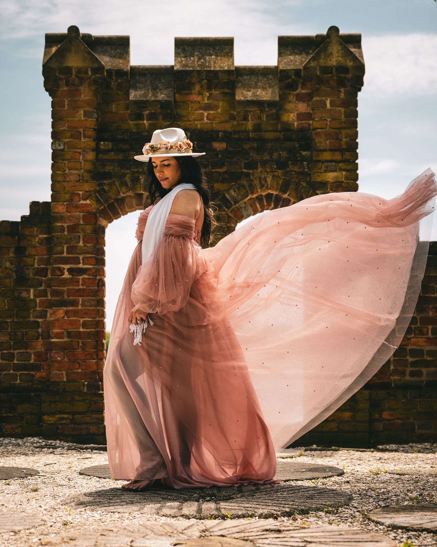 Woman in flowing pink tulle gown and floral hat poses gracefully before rustic brick ruins on a breezy day.