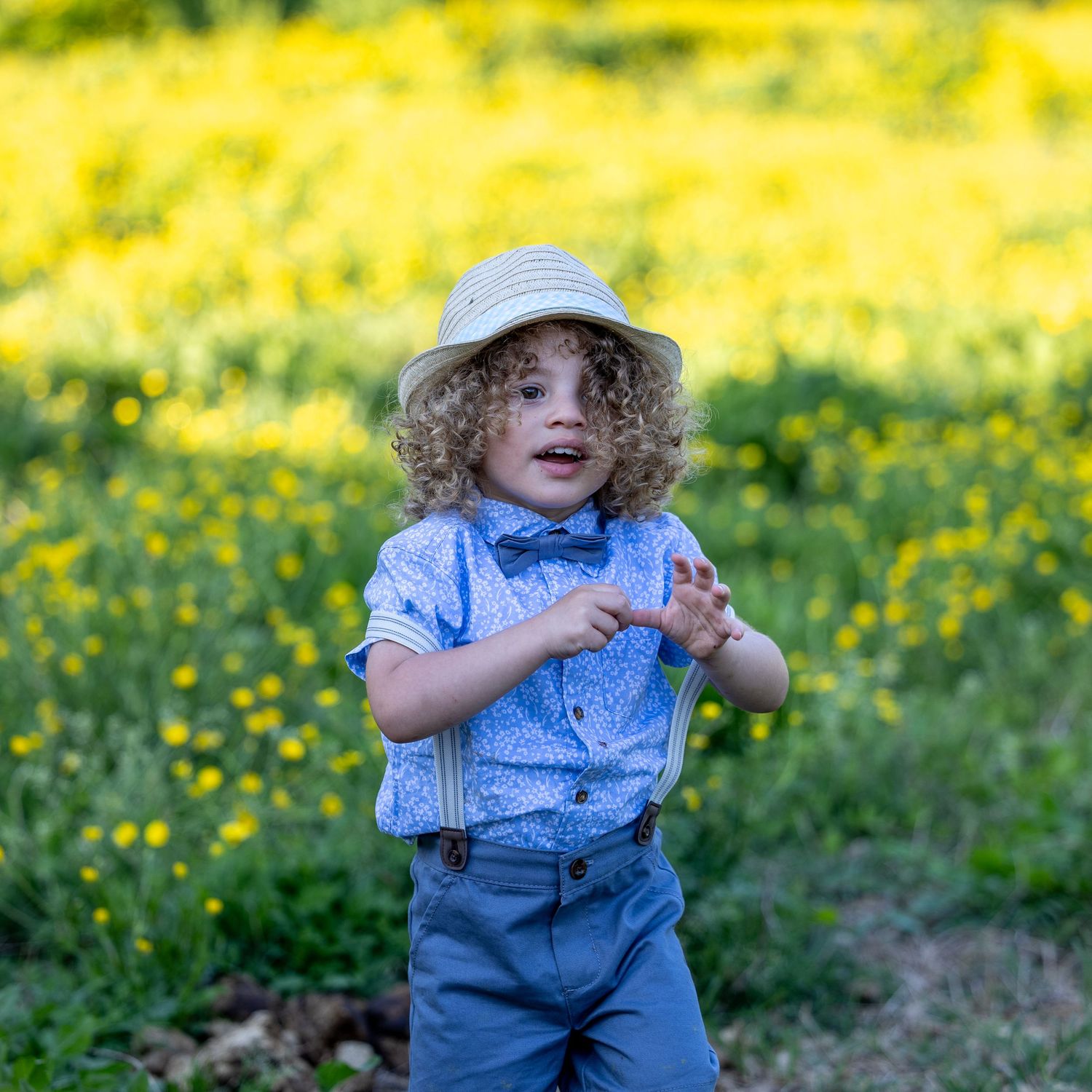 Curly-haired toddler boy in blue shirt, suspenders, and straw hat posing in a yellow wildflower field.