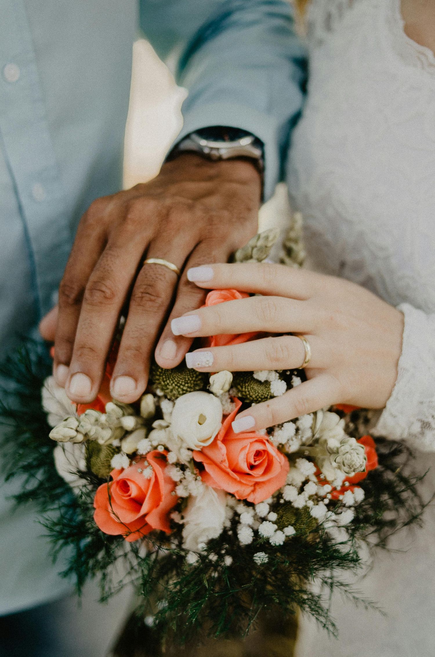 Newlywed couple displaying wedding rings on hands resting over a bouquet of coral roses.