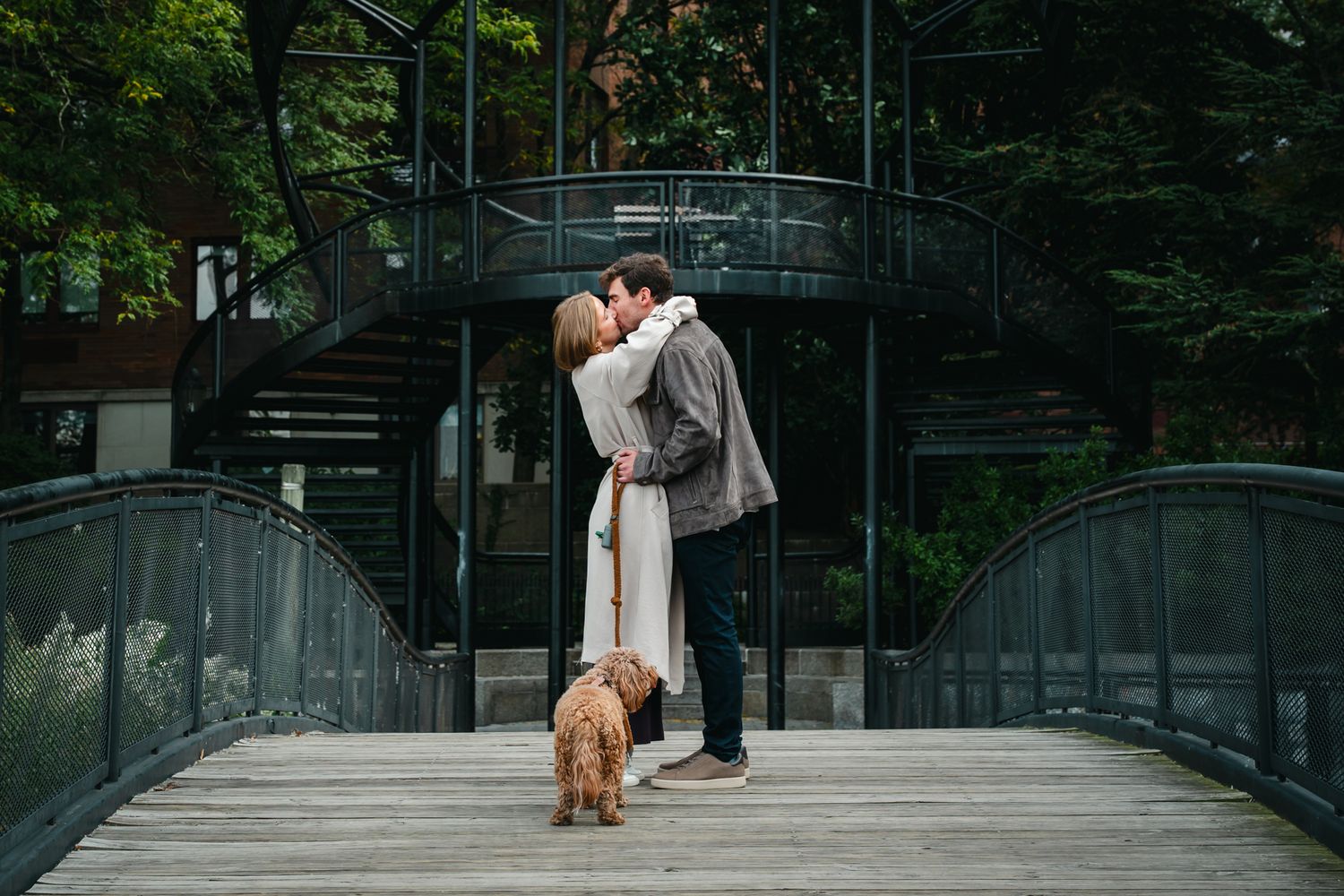 A couple embraces in a romantic moment on a modern glass bridge surrounded by lush forest scenery.
