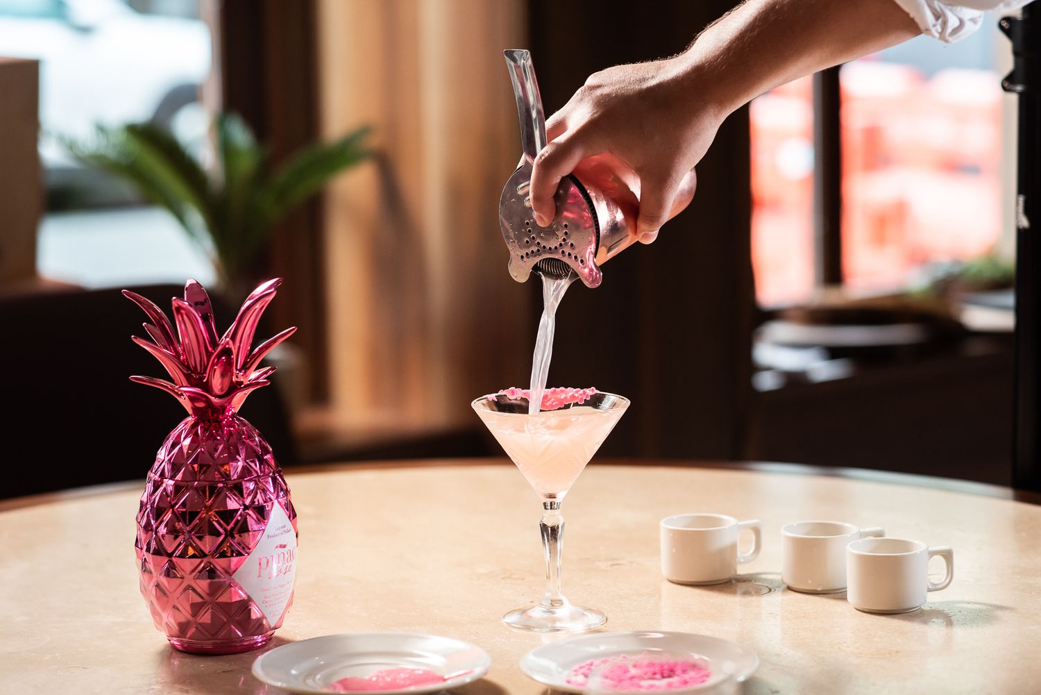 Pink cocktails being poured into glasses with decorative pineapple garnishes on a dark wooden bar.