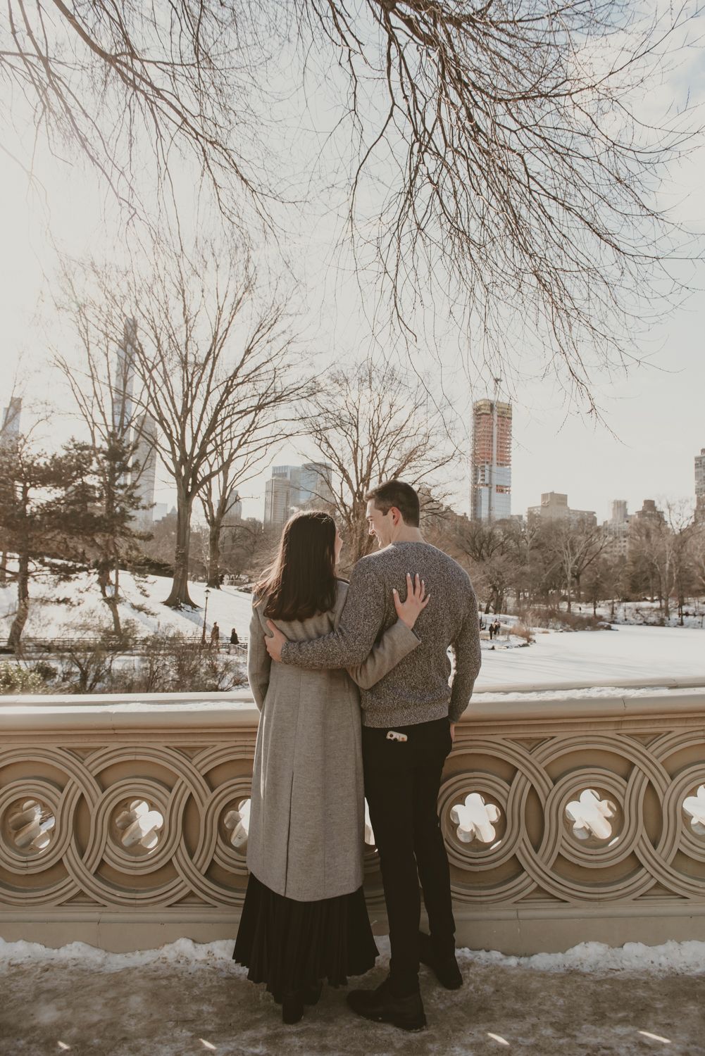 Couple embracing by an ornate stone railing in a snowy winter park with city skyline in background.