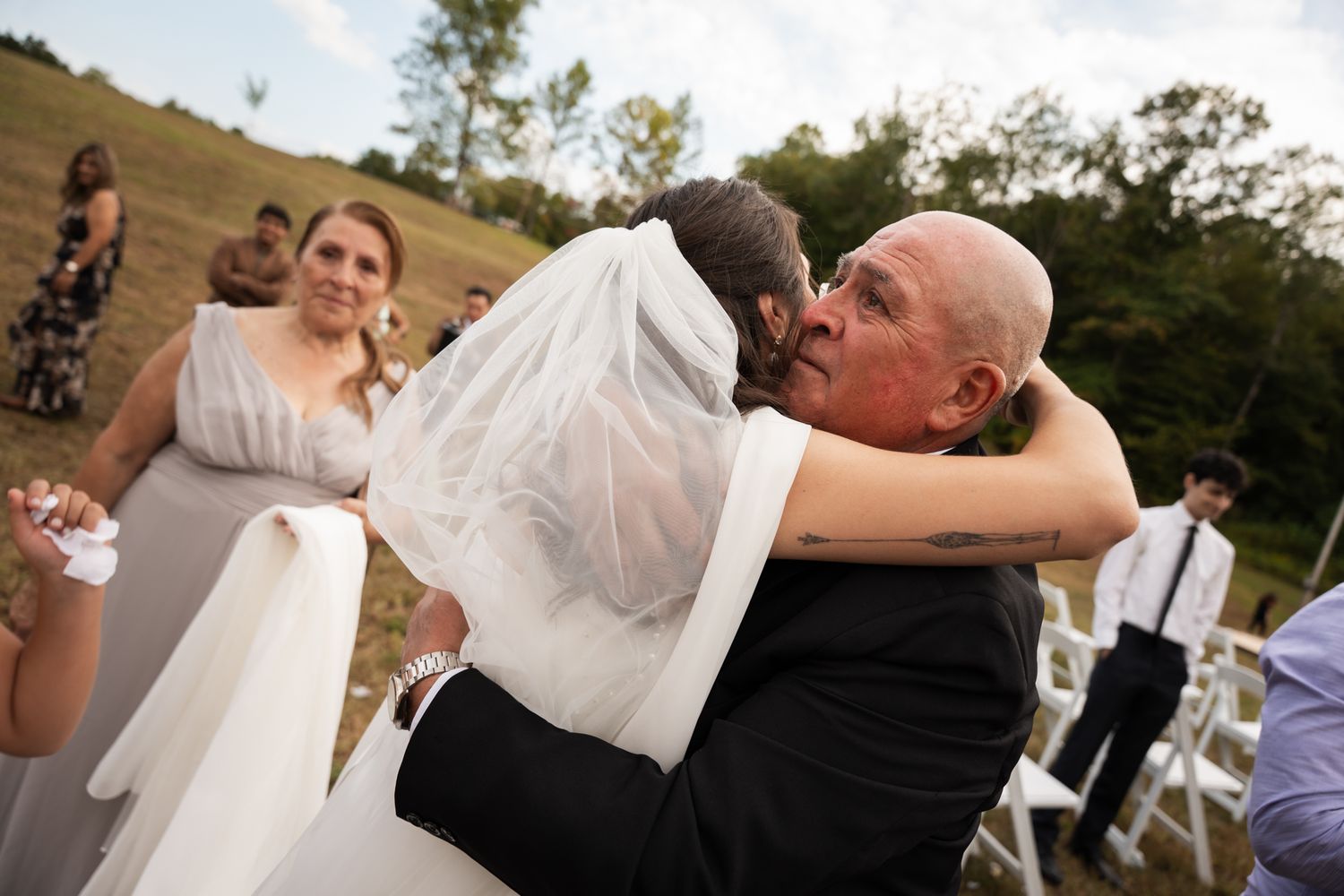 Emotional embrace during outdoor wedding ceremony on hillside with guests looking on.