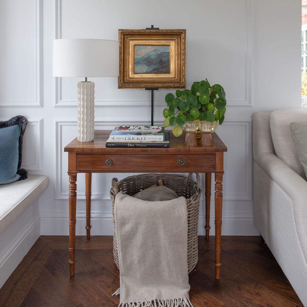 Elegant vintage wooden side table with turned legs displayed against white panelled wall in a bright living room. Antique oil painting.