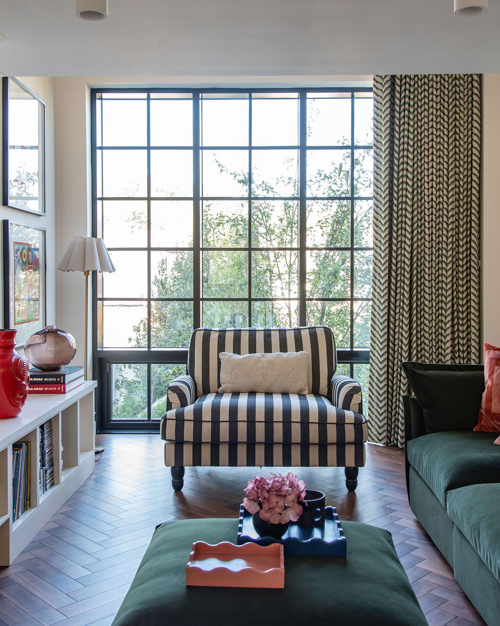 Modern living room with green velvet sofa, striped curtains, and large windows overlooking lush greenery outside.