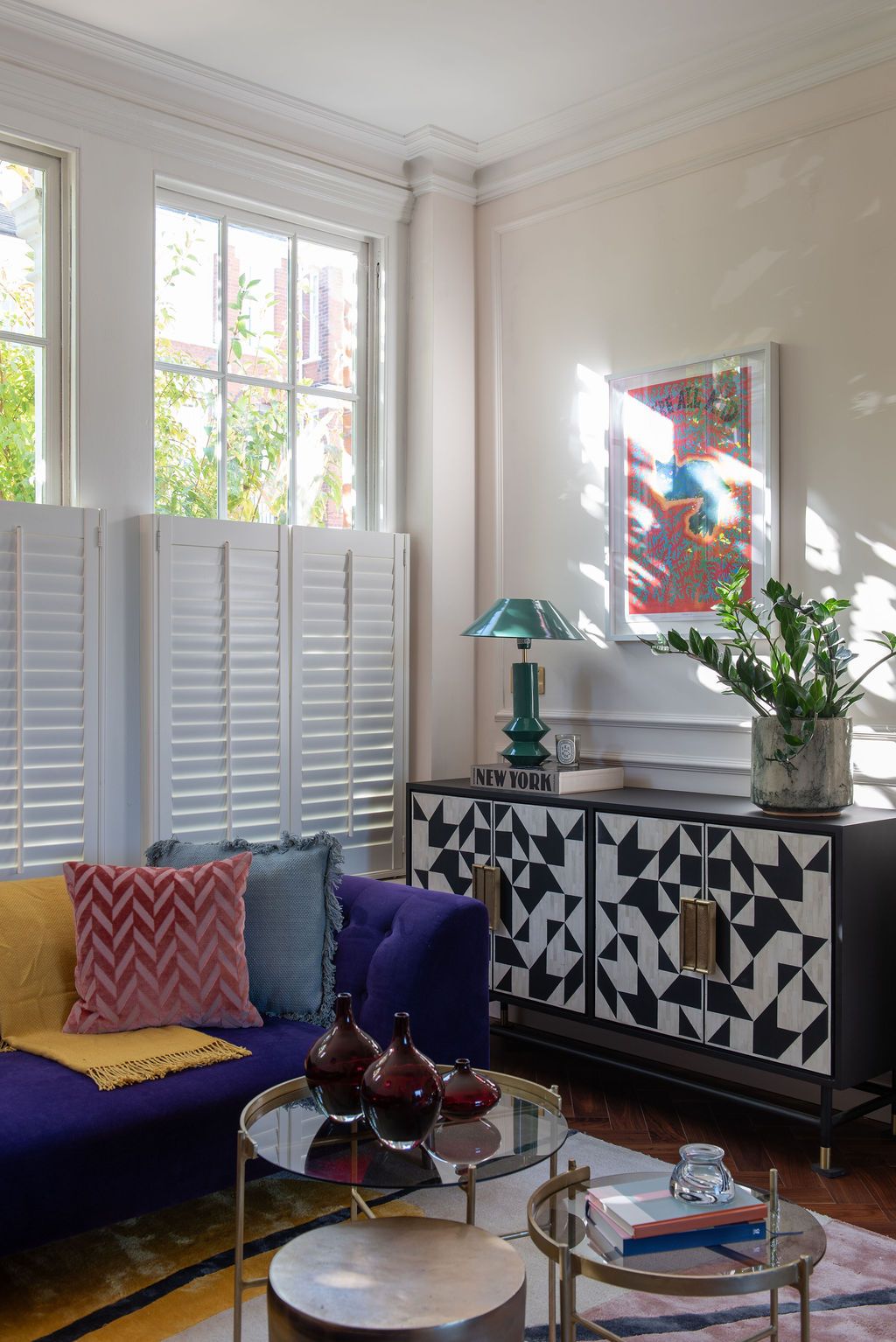 Bright sunroom with purple sofa, patterned throw pillows, white shutters, and black and white geometric side board with green table lamp