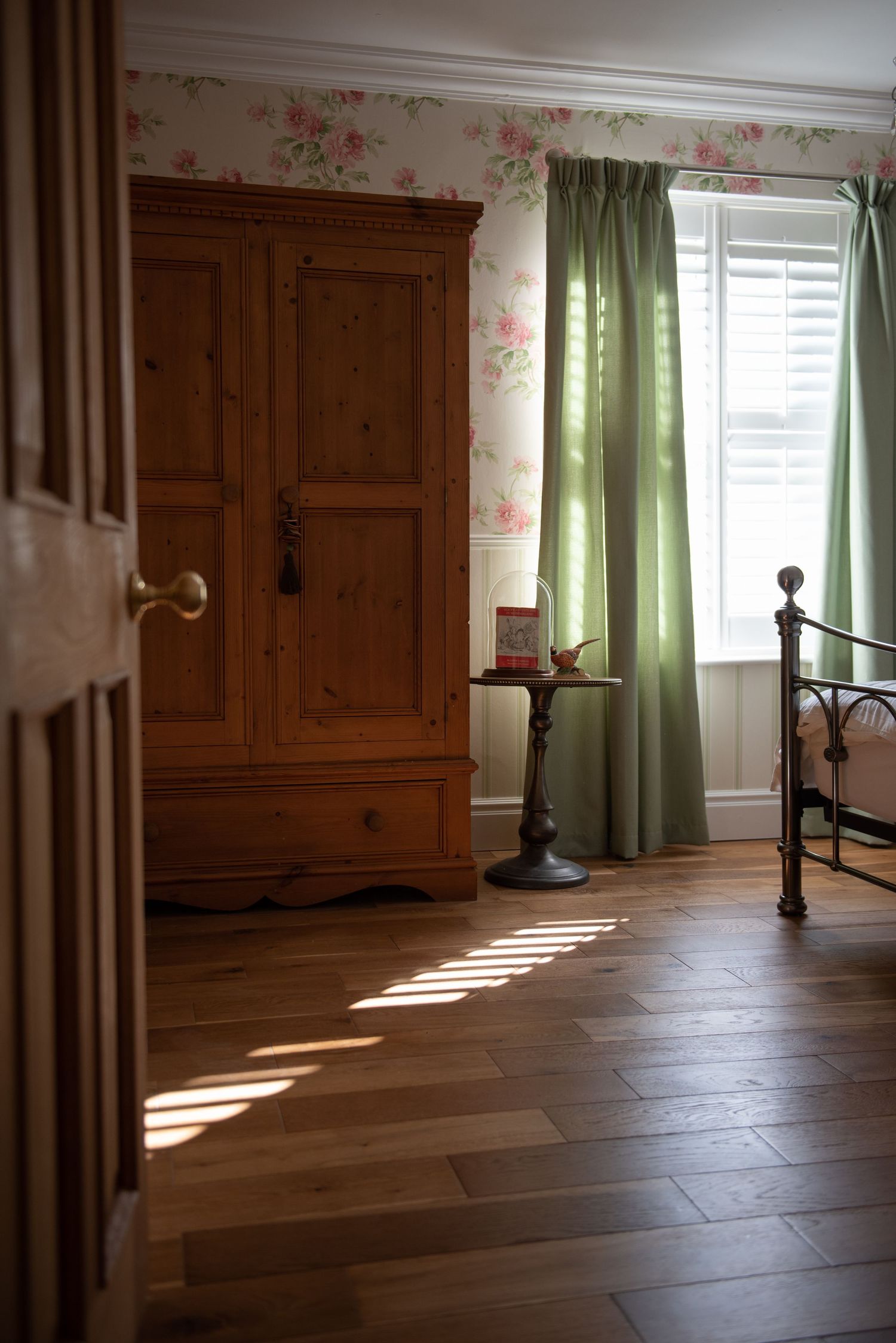 Sunlight streams through green curtains into a room with wooden furniture and floral wallpaper, creating dramatic shadows.