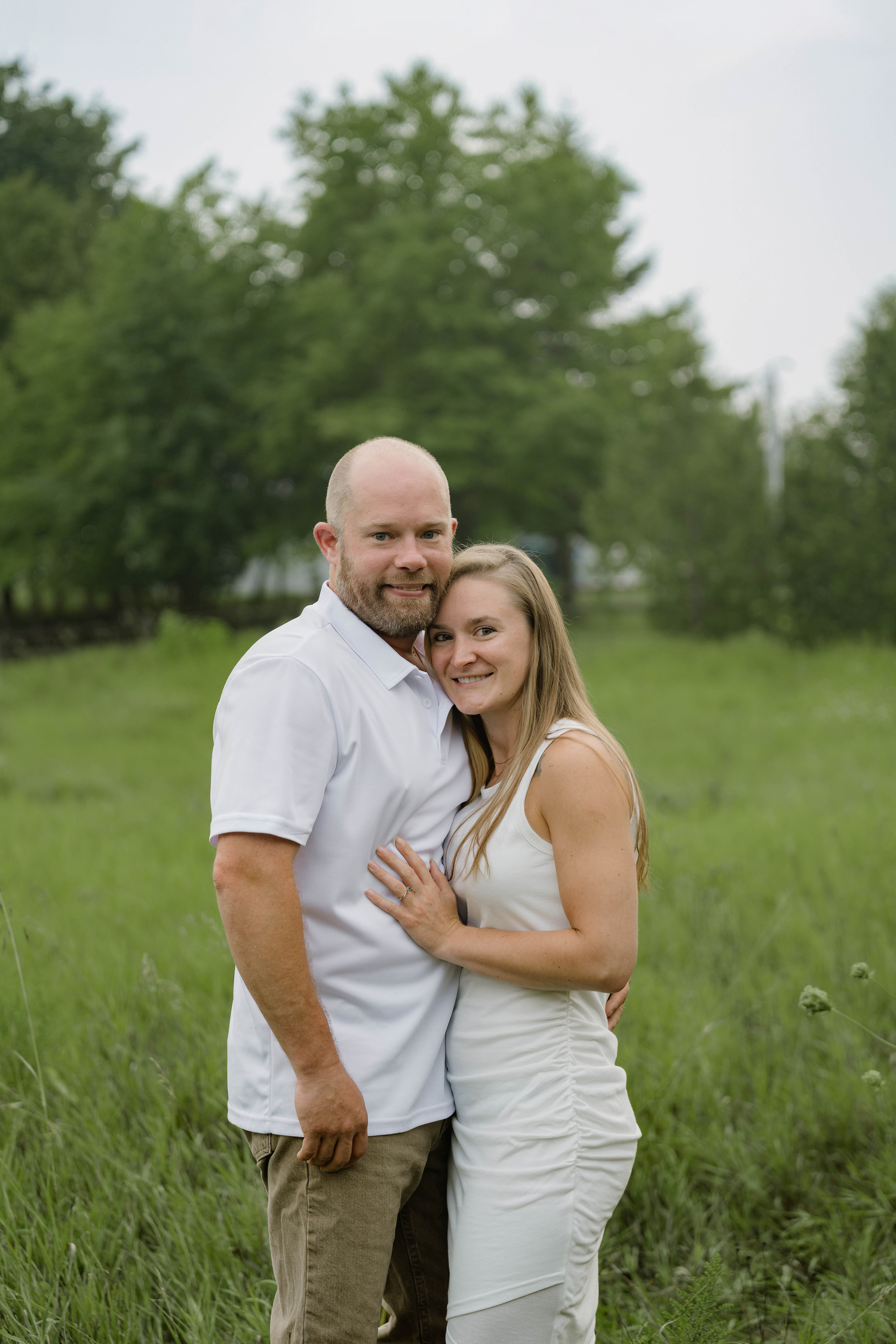 A smiling couple poses together in a green field, the woman resting her head on the mans chest.