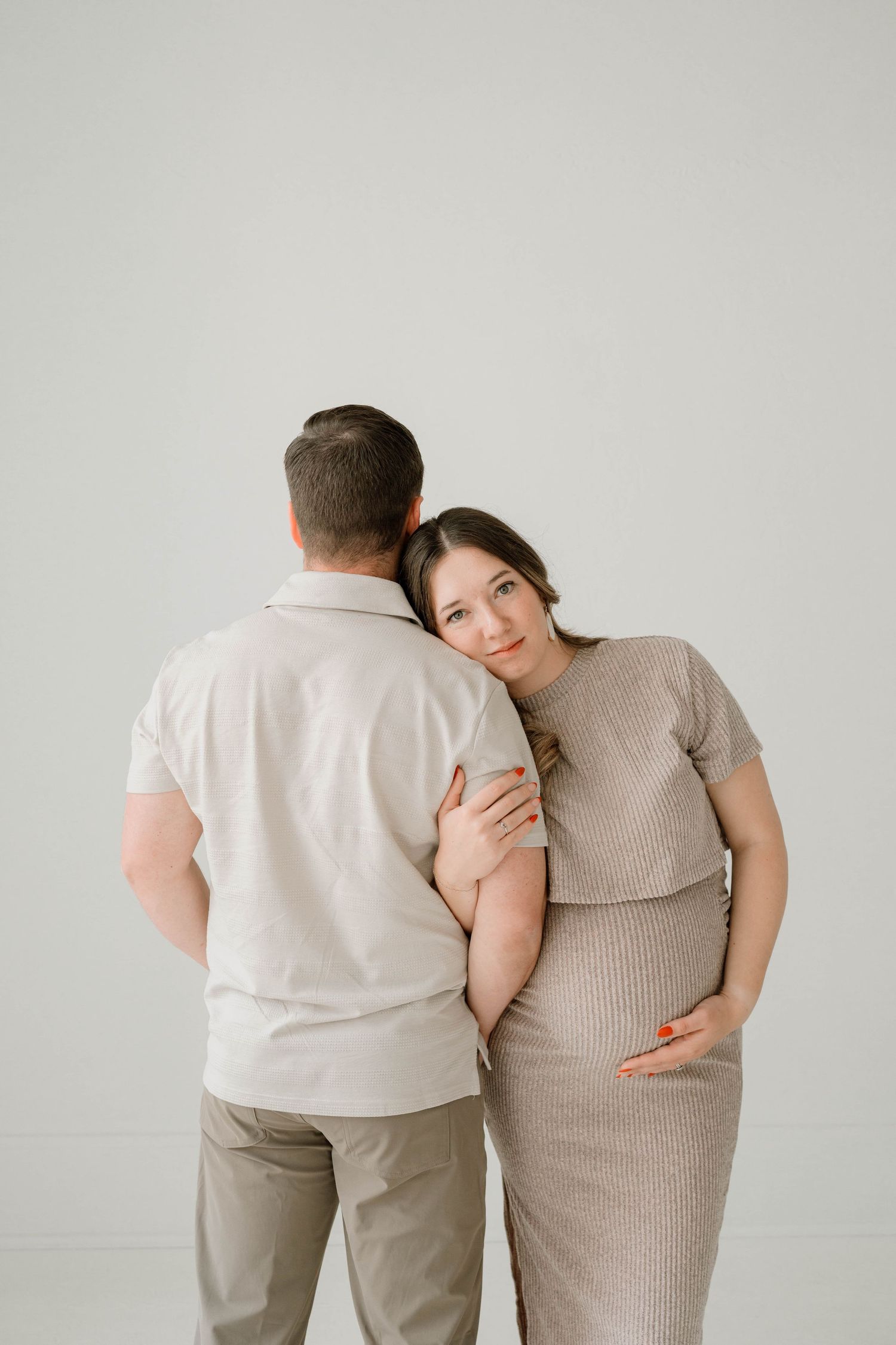 Pregnant woman leaning on partner's back, hand on bump, looking at camera softly.