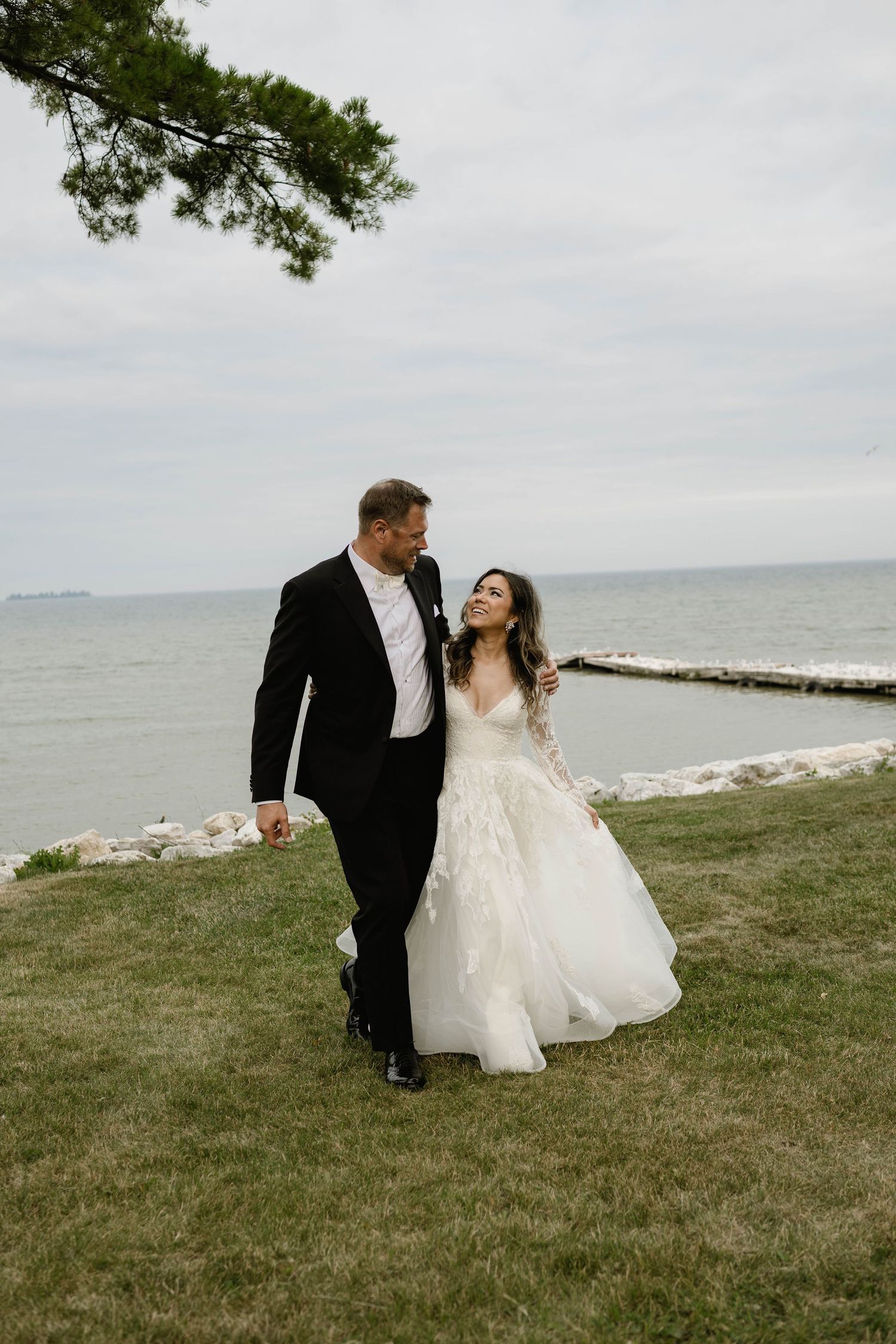 Bride and groom walking hand in hand along grassy waterfront, gazing lovingly at each other.