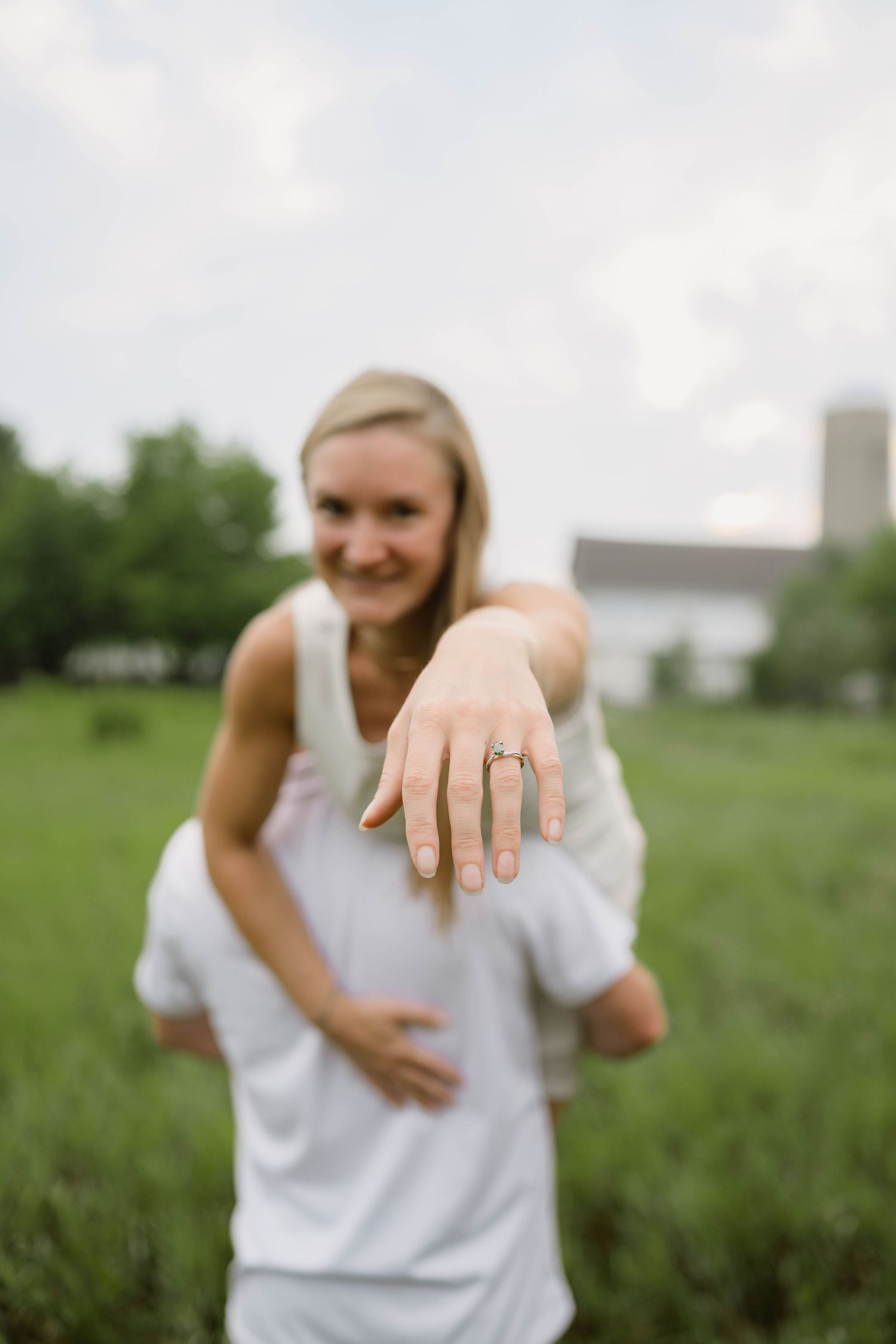 Engaged woman showing off her ring while being carried by her partner in a green field.