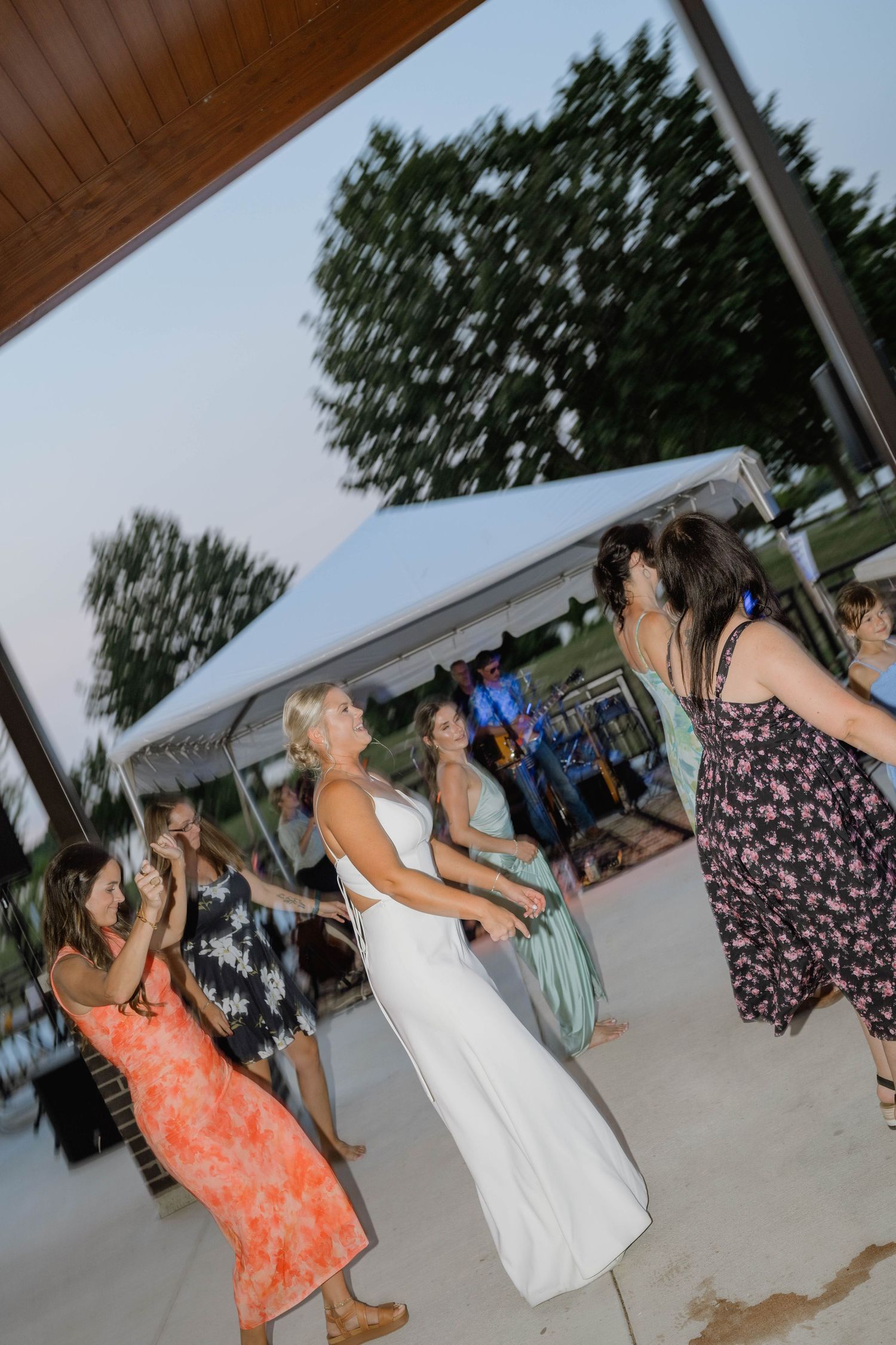 Bride in white dress dances energetically with guests at outdoor wedding reception under evening sky.