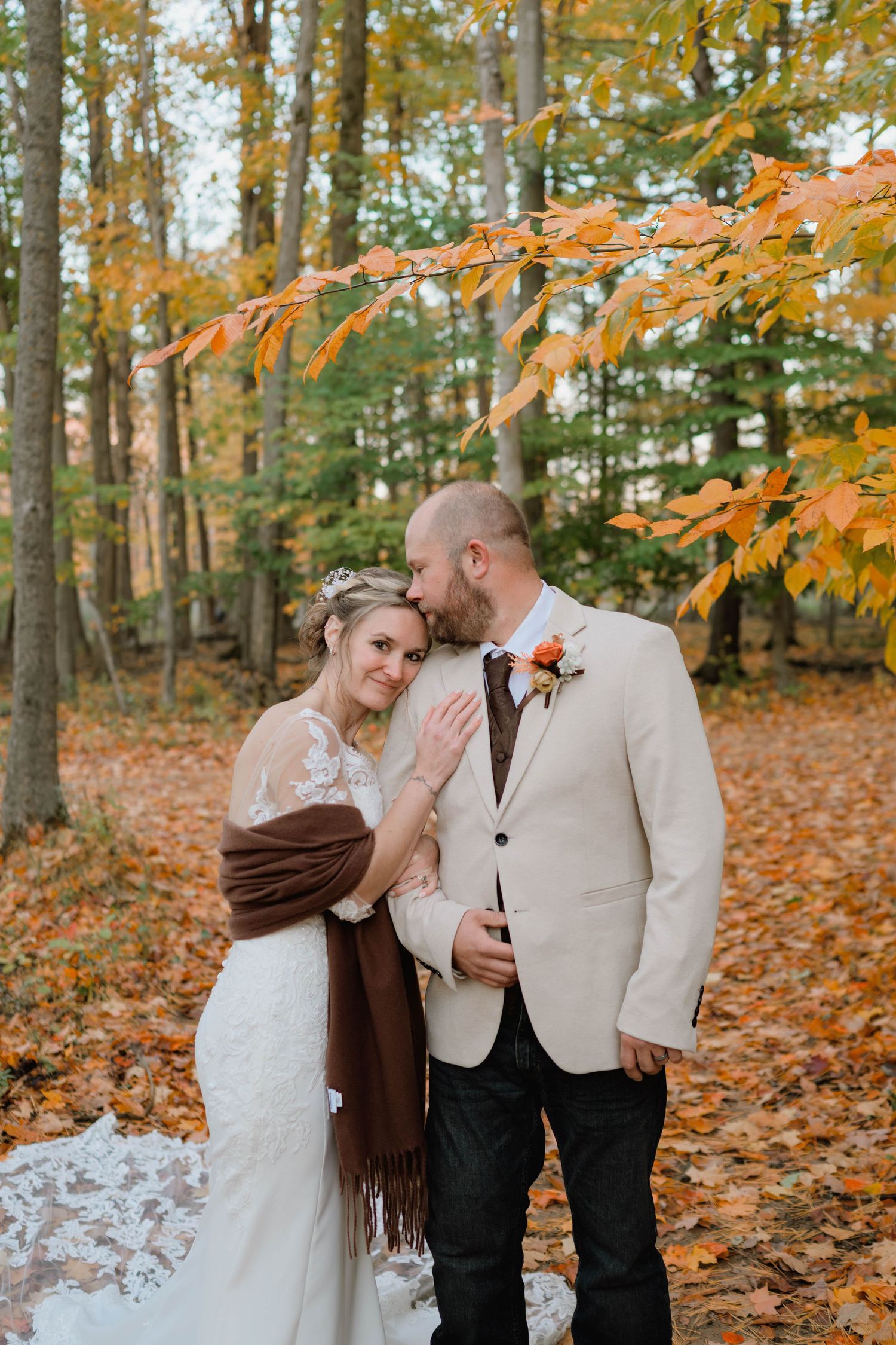 Bride in a lace wedding gown and burgundy shawl embraces her groom in a cream blazer as he kisses her forehead among autumn foliage.