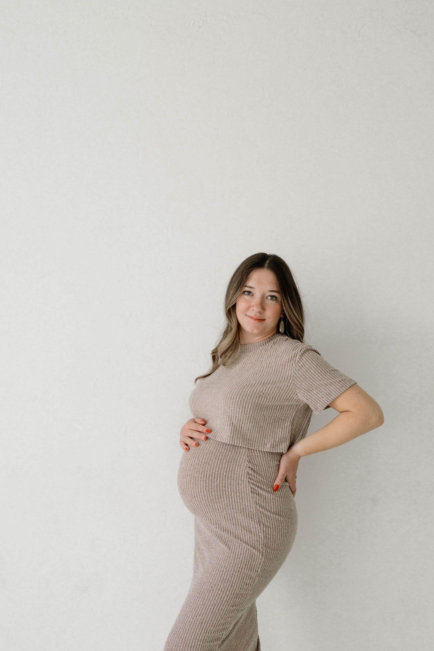 Smiling pregnant woman in beige ribbed dress cradles baby bump against clean white studio wall.