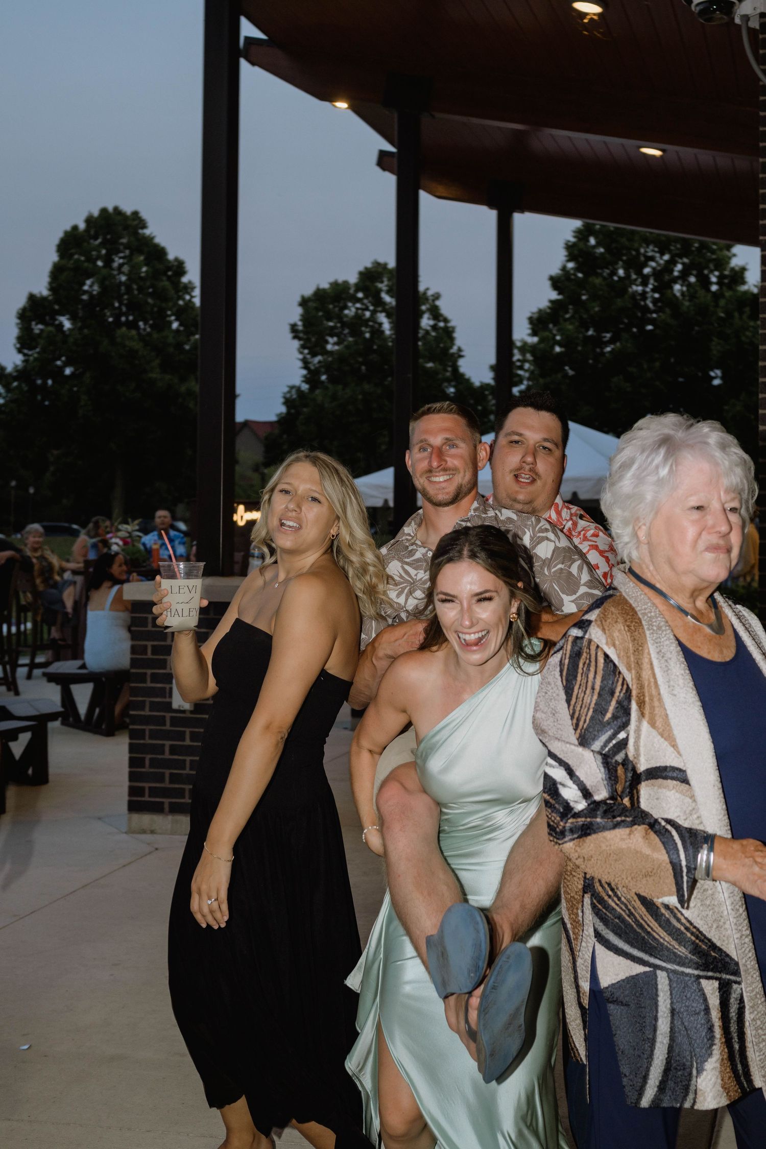 Excited wedding guests laugh and pose together at outdoor evening wedding reception under pavilion.