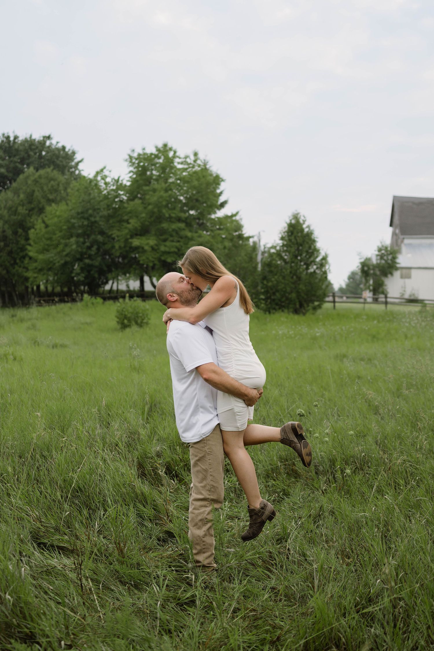 Man lifting and kissing woman in white dress in a green field with trees and barn in background.