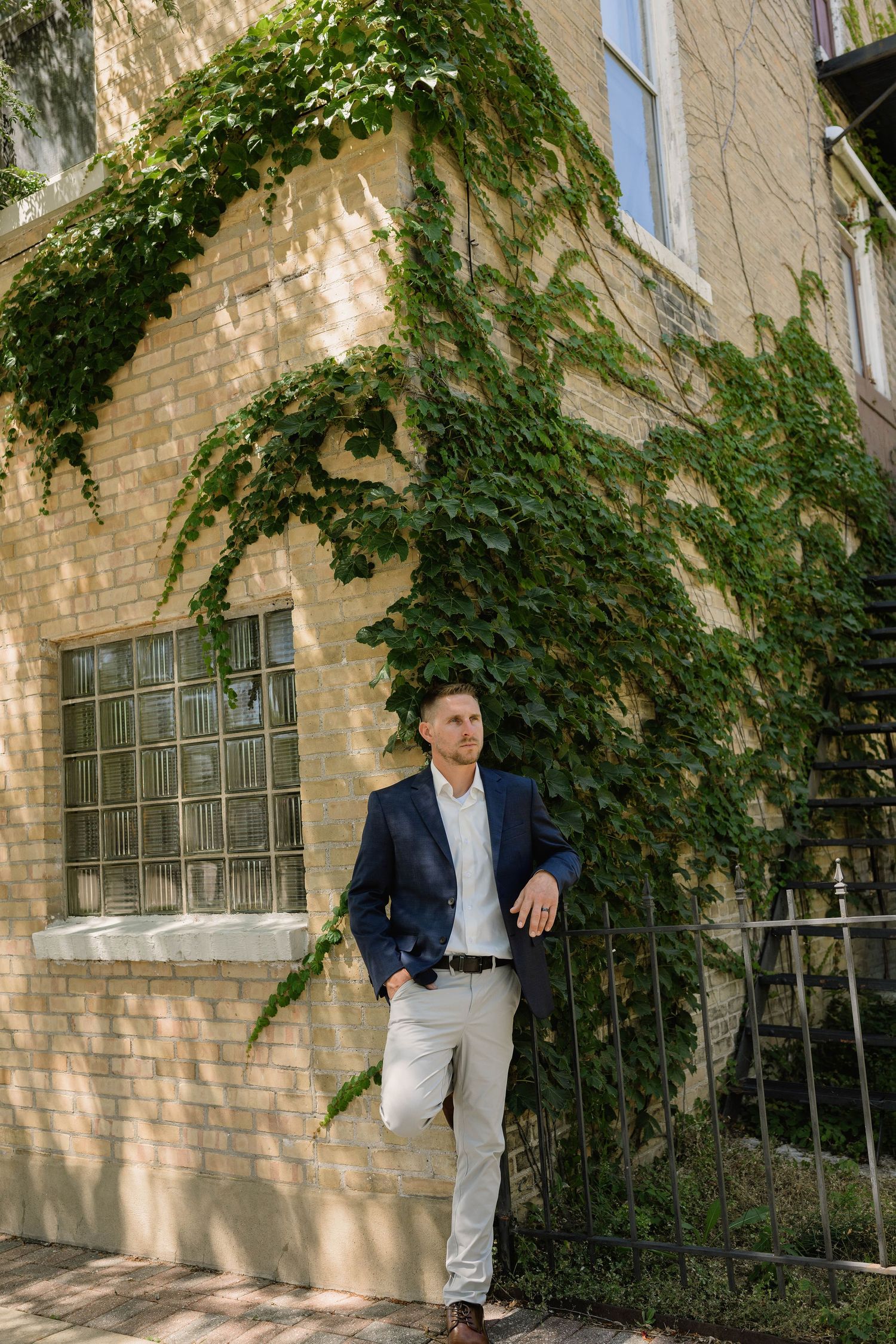 Groom in navy suit leaning against iron fence by ivy-covered building.