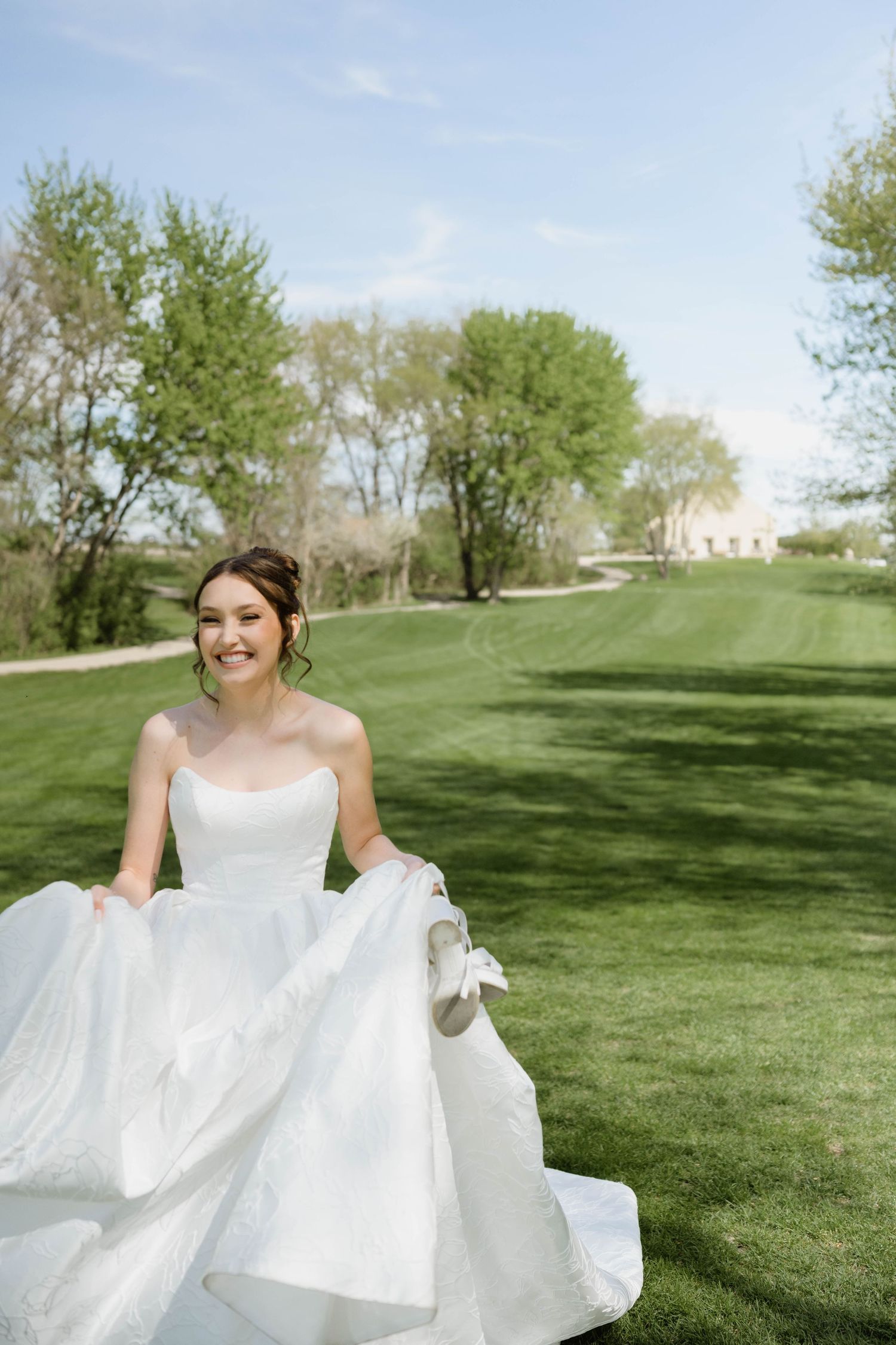 Smiling bride in a strapless white ball gown lifts her skirt while walking on a sunny green lawn.