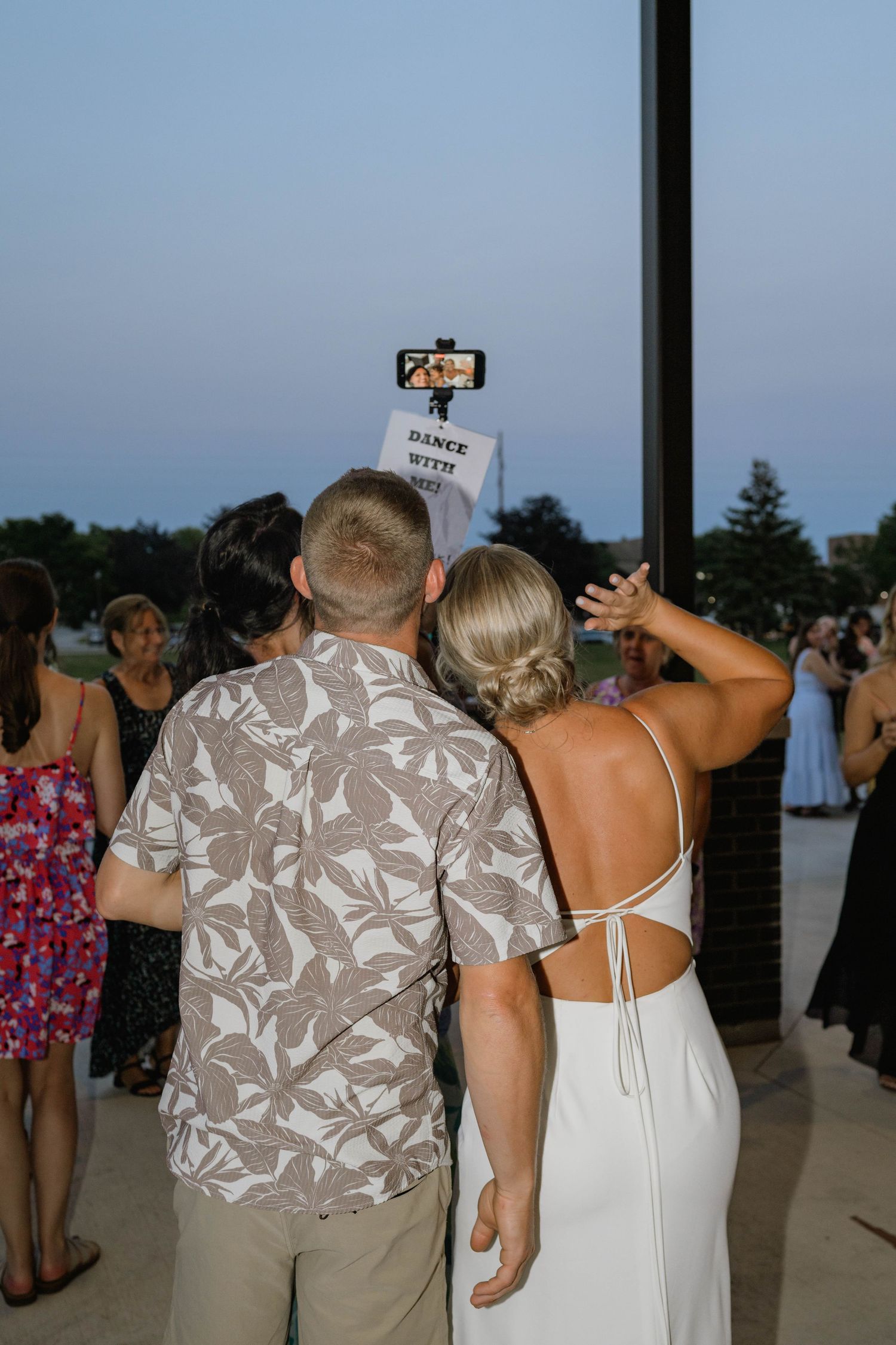 Bride in backless white gown takes selfie with guest using phone on selfie stick at wedding.