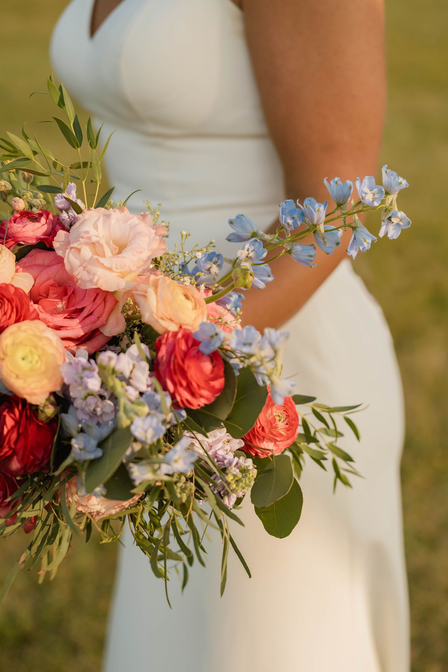 Bride in white gown holds lush colorful bouquet with pink, coral, and blue blooms.