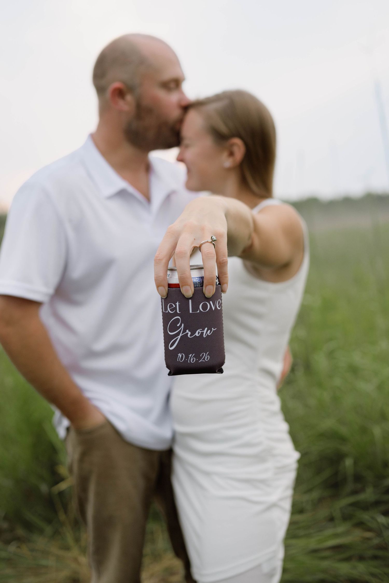 Couple kissing while showing a koozie reading Let Love Grow with their wedding date in a field.