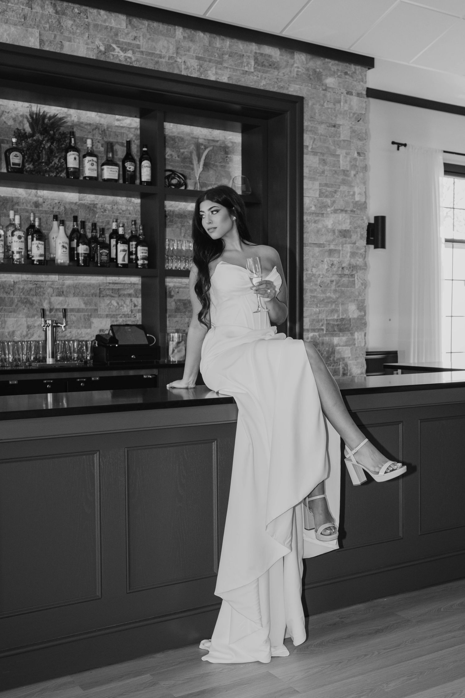 Black and white photo of bride in elegant white gown perched on bar counter with champagne glass.