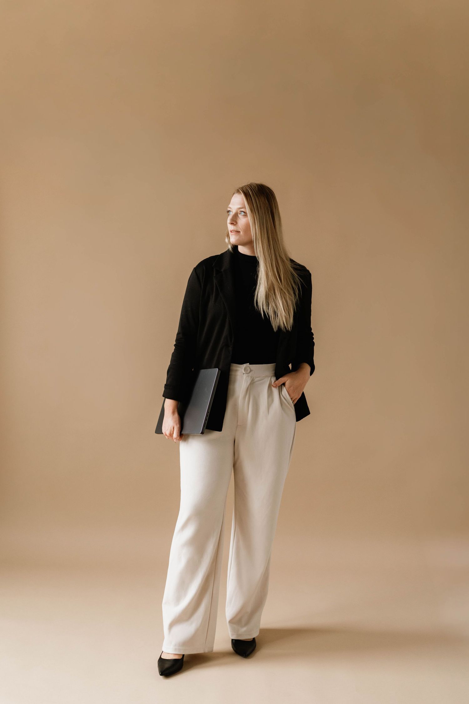 Professional woman in black blazer and white wide-leg trousers holding a laptop against beige background.