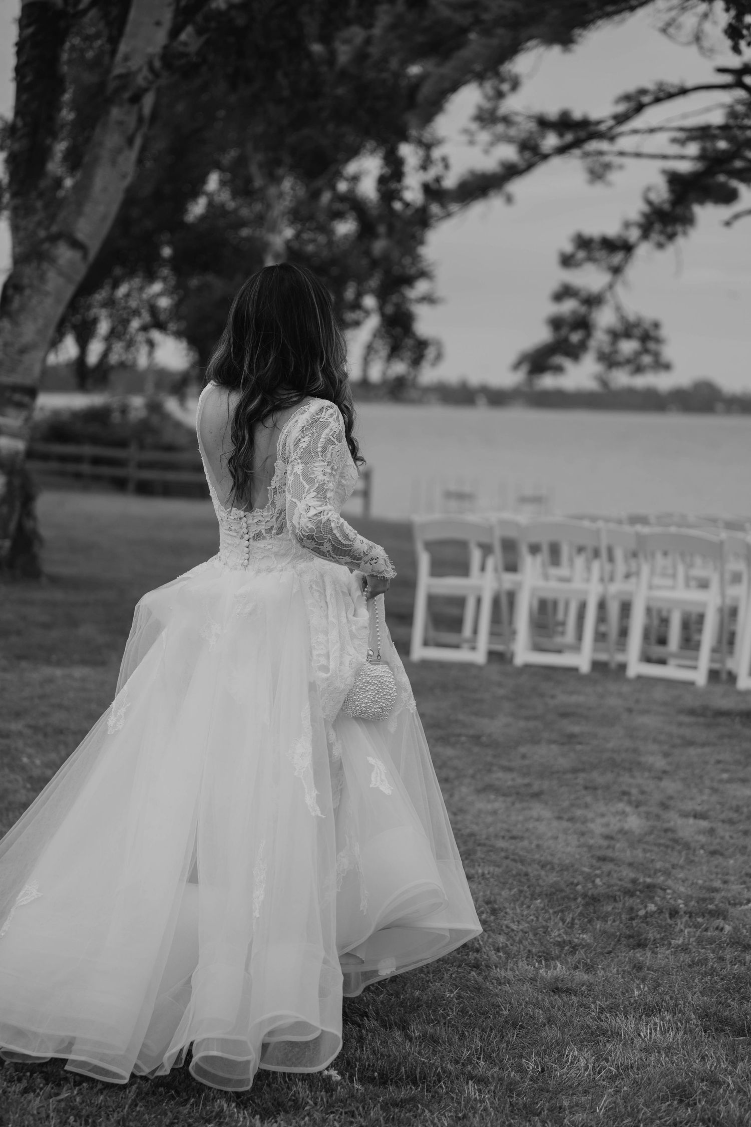 Black and white rear view of bride in lace wedding gown near lakeside ceremony chairs.