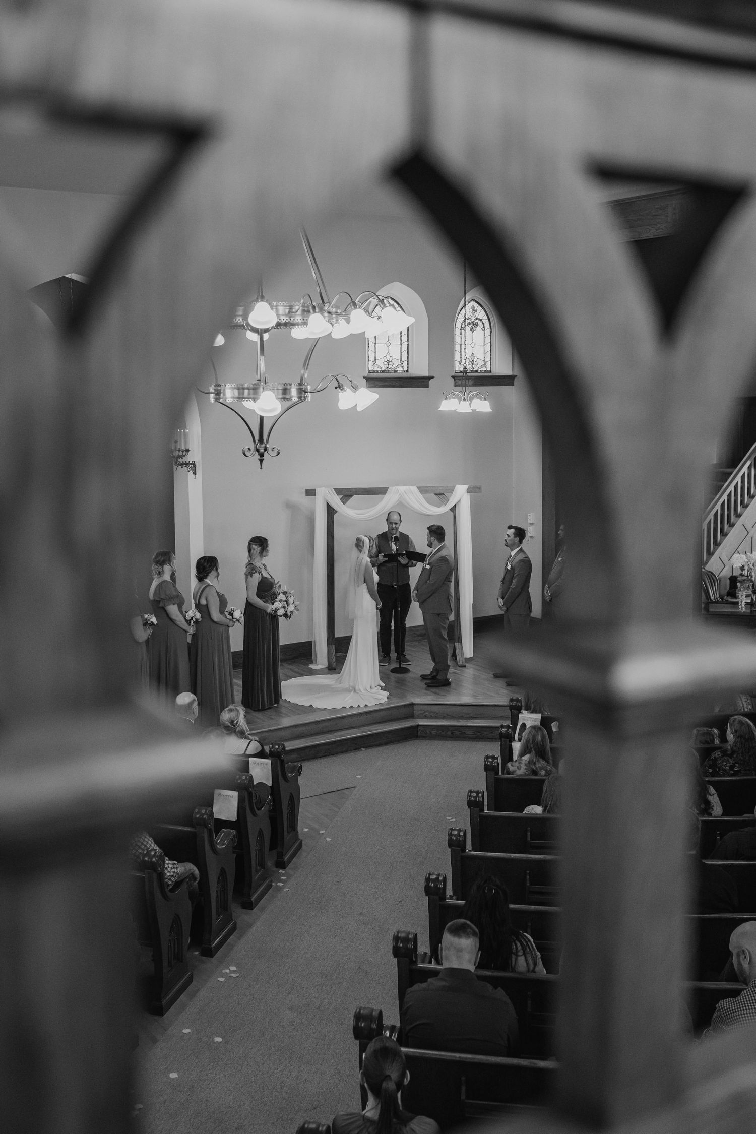 Black and white photo of a bride and groom at the altar during a church wedding ceremony, framed by ornate wooden pews in the foreground.