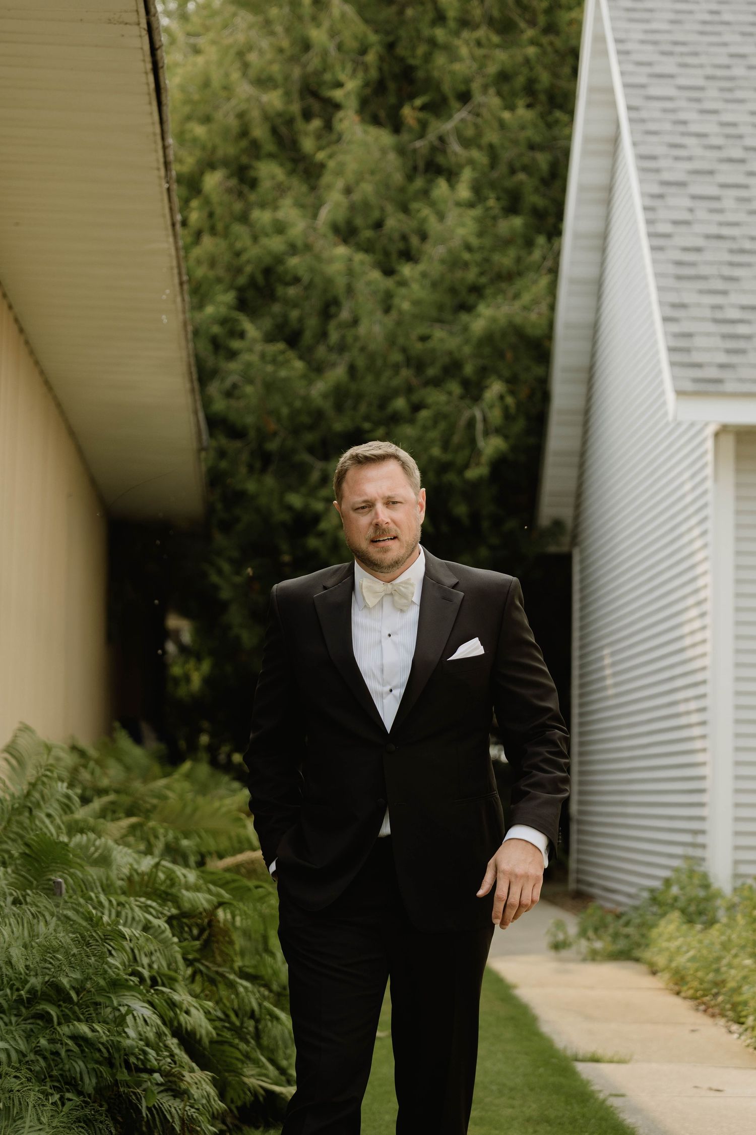 Dapper groom in a black tuxedo with white pocket square stands confidently between two buildings surrounded by greenery.