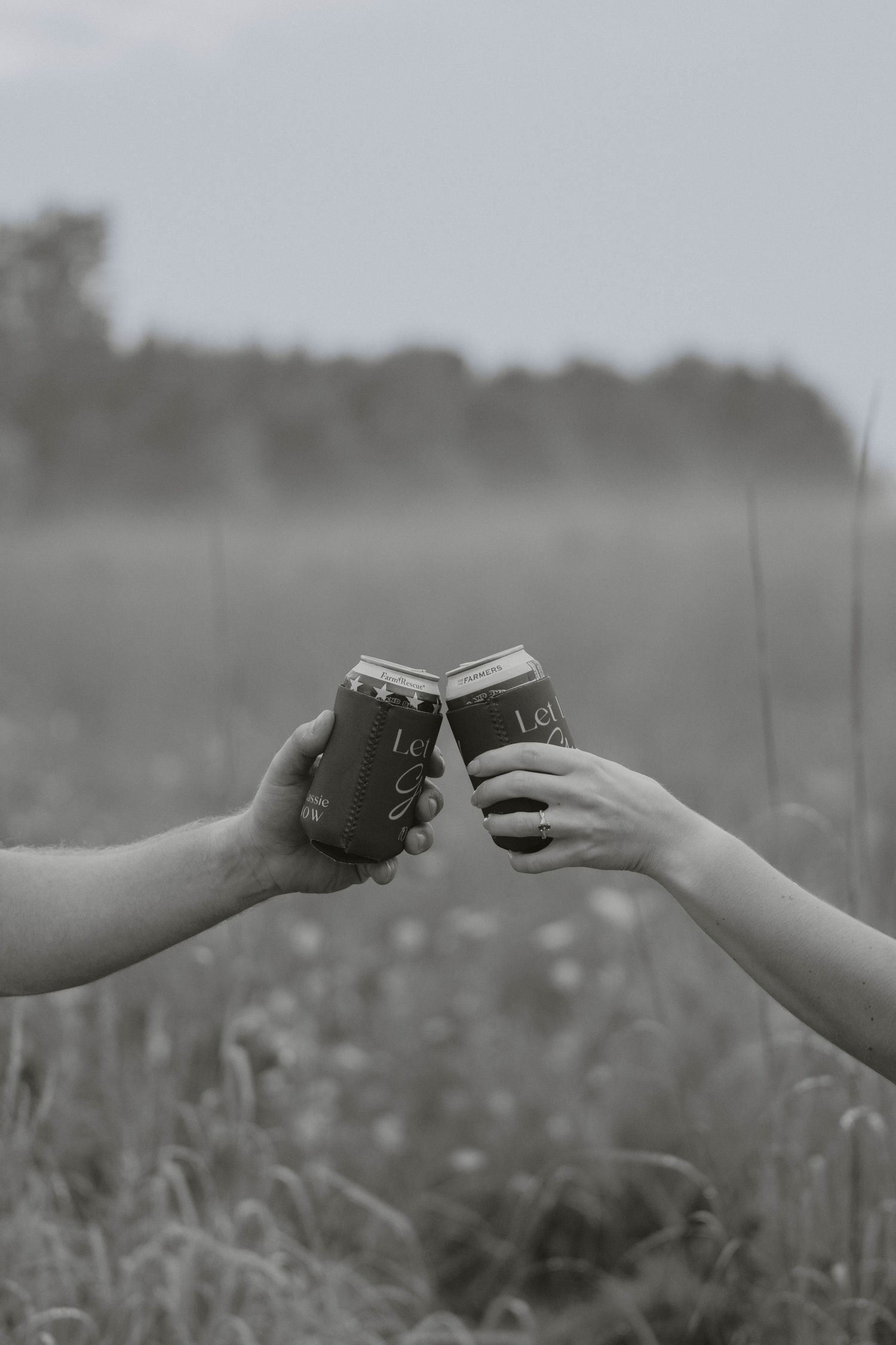Black and white close-up of two hands clinking mason jar cups in a grassy outdoor field.