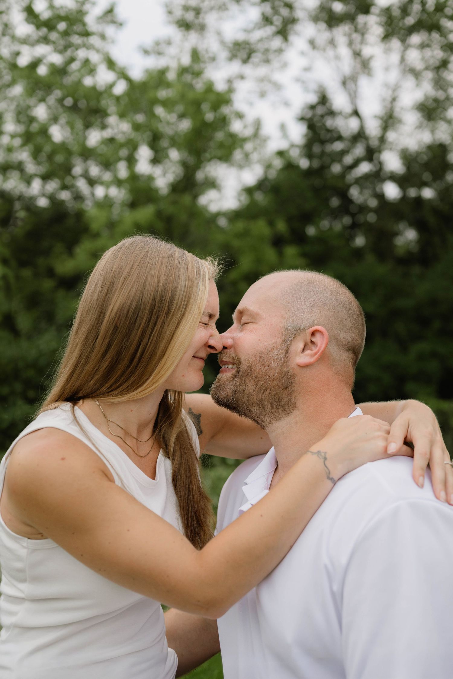 A couple shares a tender nose-to-nose moment while smiling in a lush green outdoor setting.