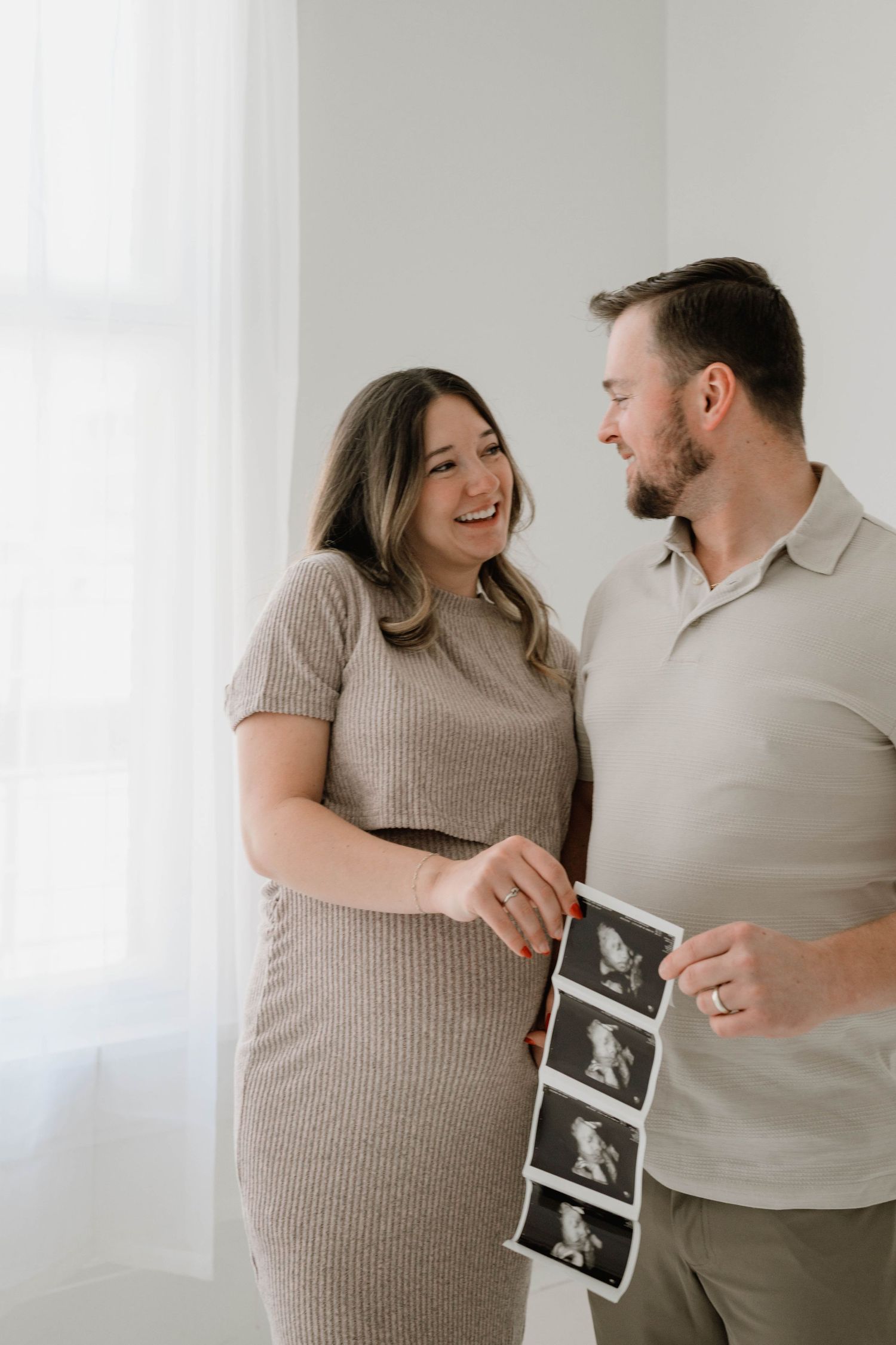 Happy expecting couple laughs together while holding ultrasound photos in bright windowed studio.