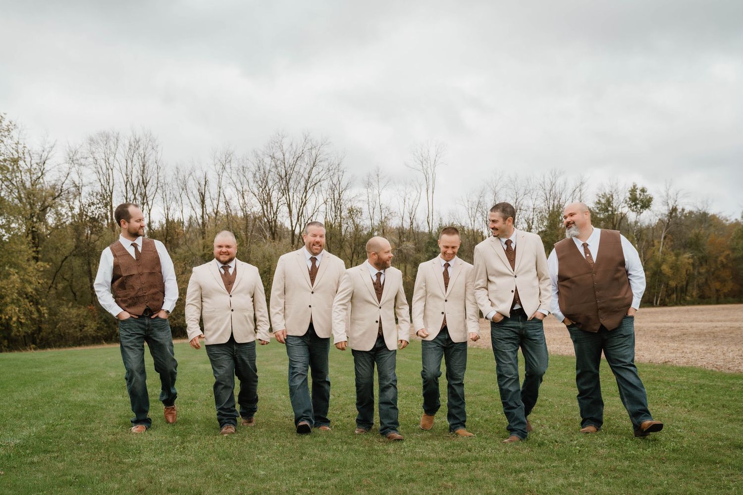 Seven groomsmen in tan suit jackets and brown vests walk side by side across a green lawn with bare autumn trees in the background.