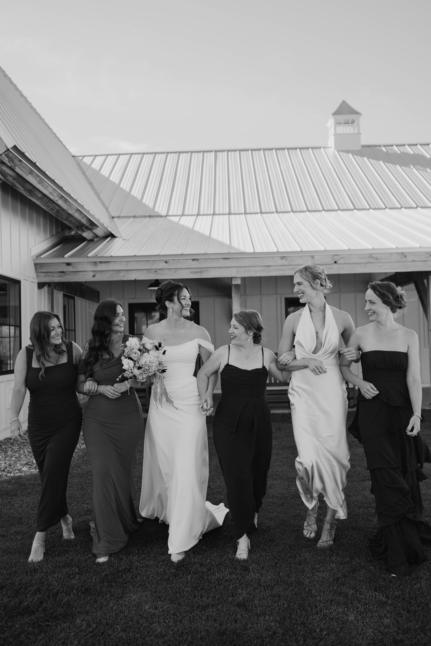Black and white photo of a bride and five bridesmaids in elegant gowns laughing together outside a barn venue.