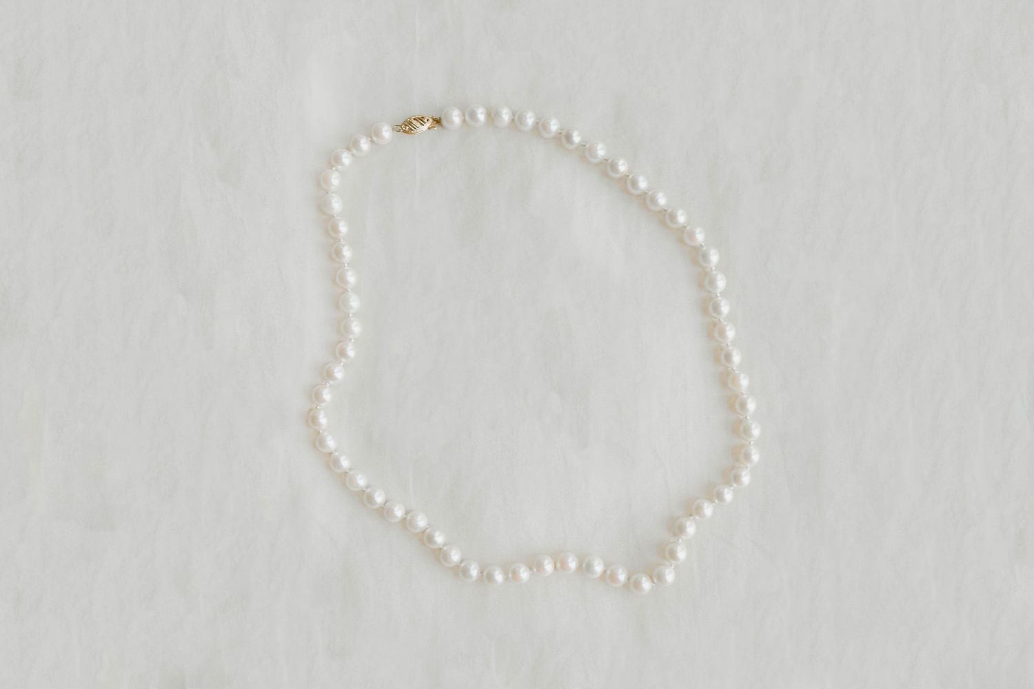 Classic pearl necklace with gold clasp laid flat on white textured fabric background.