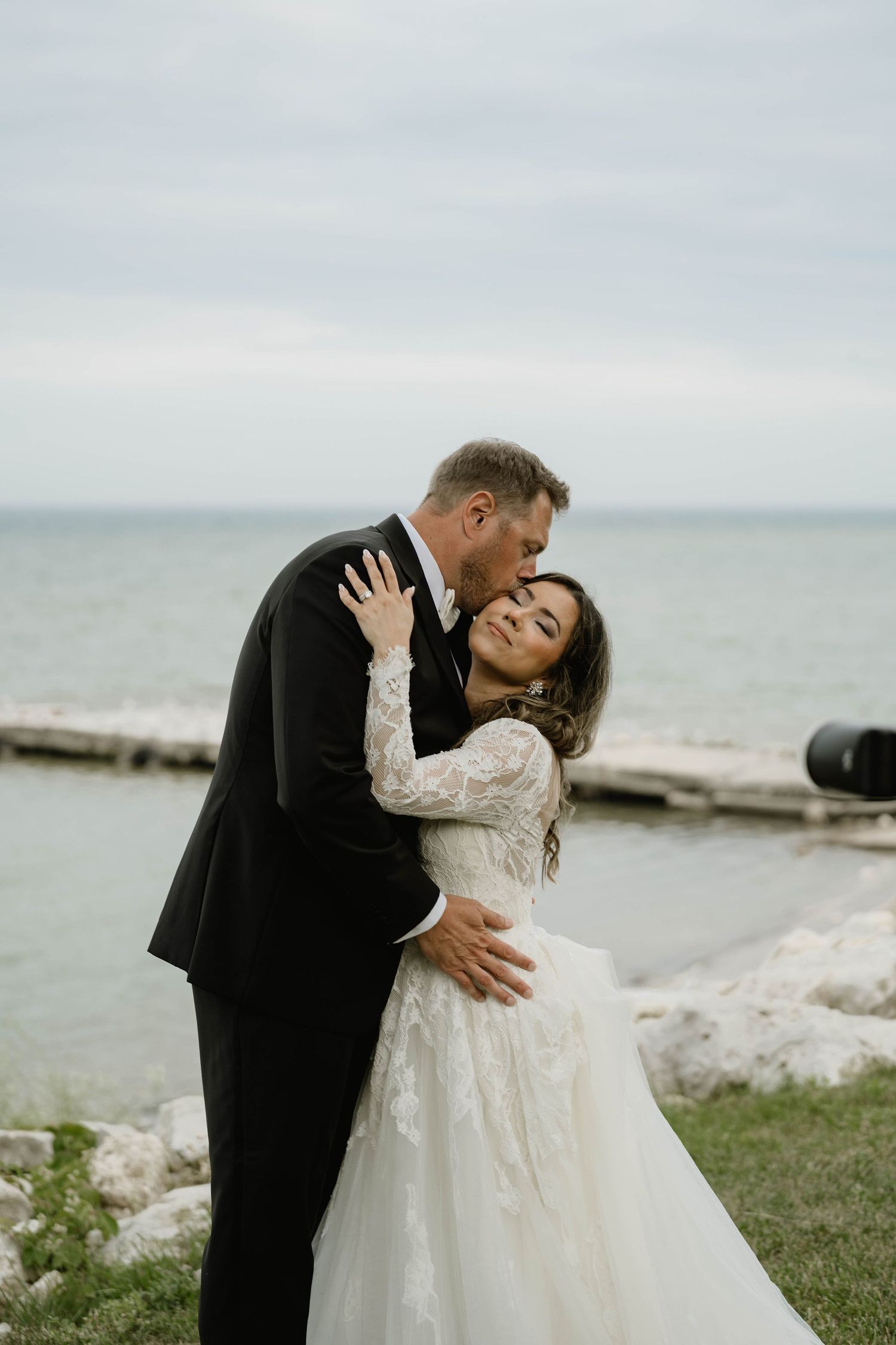 Groom in black tuxedo embracing bride in lace wedding dress near rocky shoreline on overcast day.