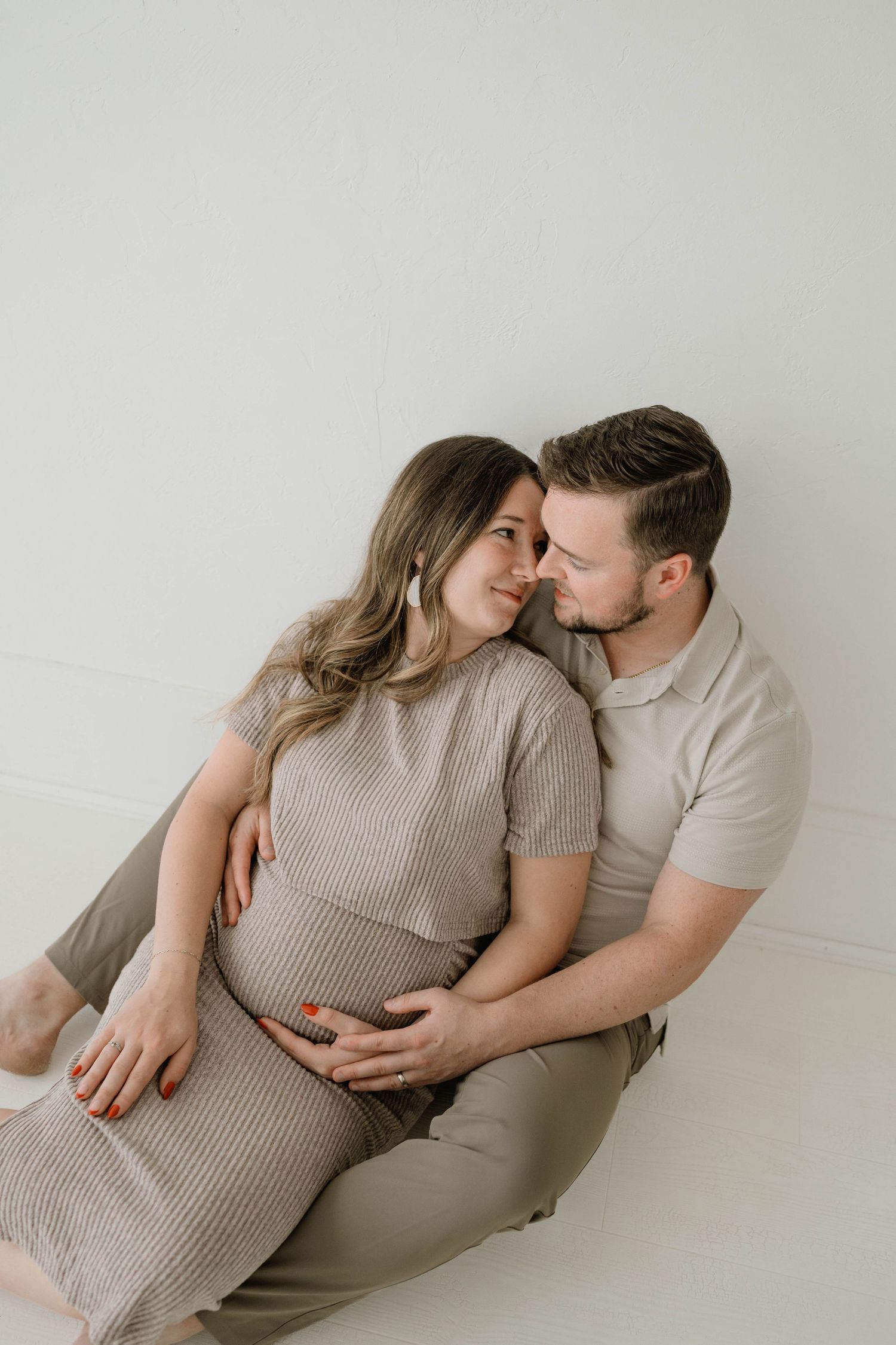Couple sharing tender moment on floor, man embracing pregnant woman from behind, both smiling at each other.