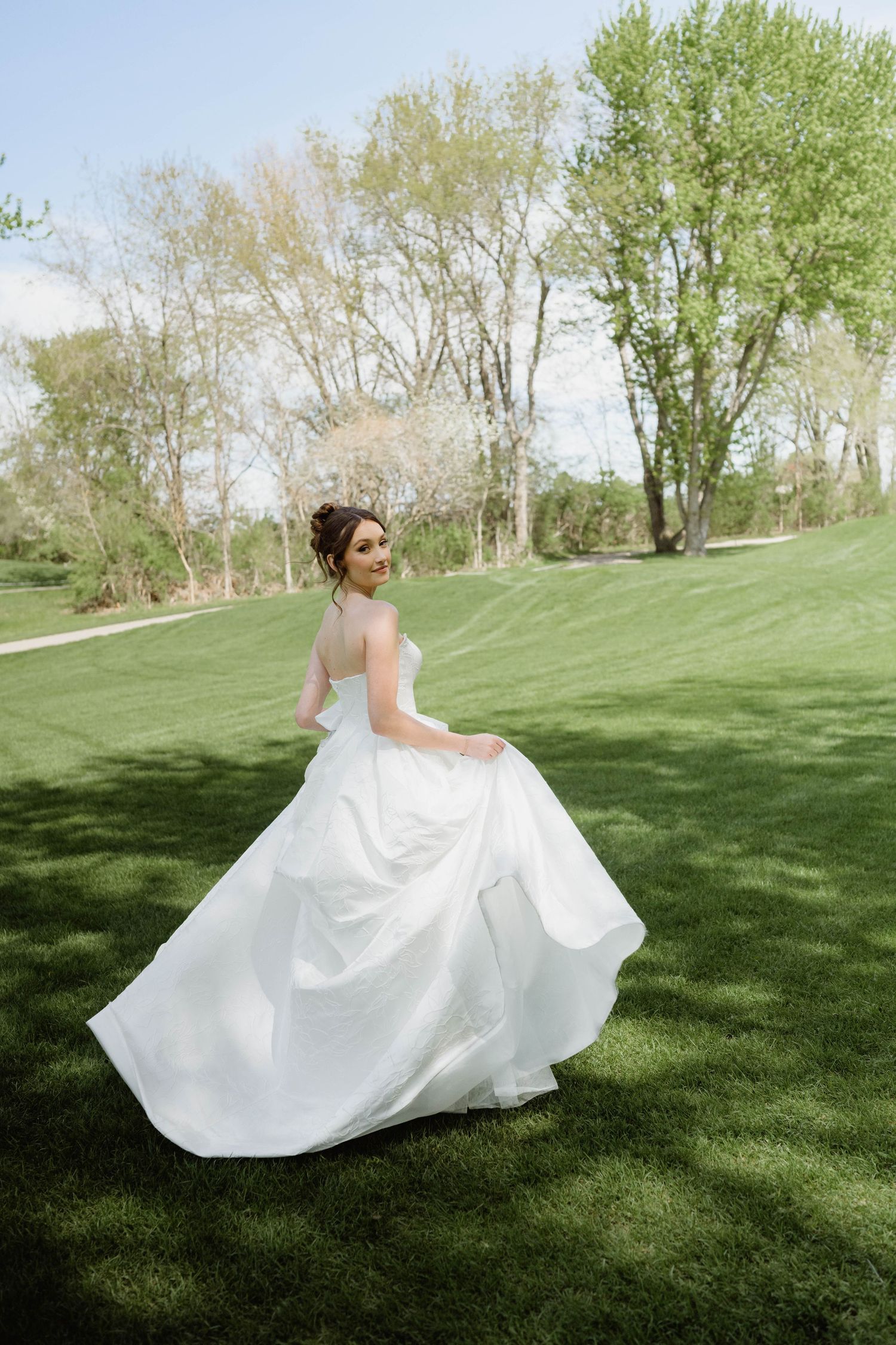Smiling bride twirls in a flowing white strapless ball gown on a green lawn surrounded by spring trees.