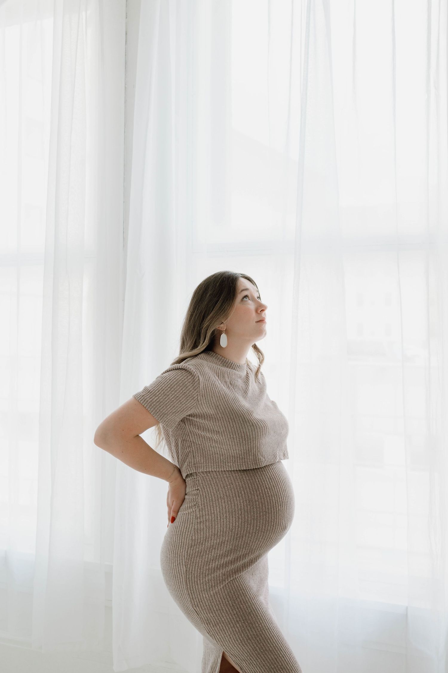 Pregnant woman in beige ribbed dress stands by white curtained window, hand on back, looking upward.