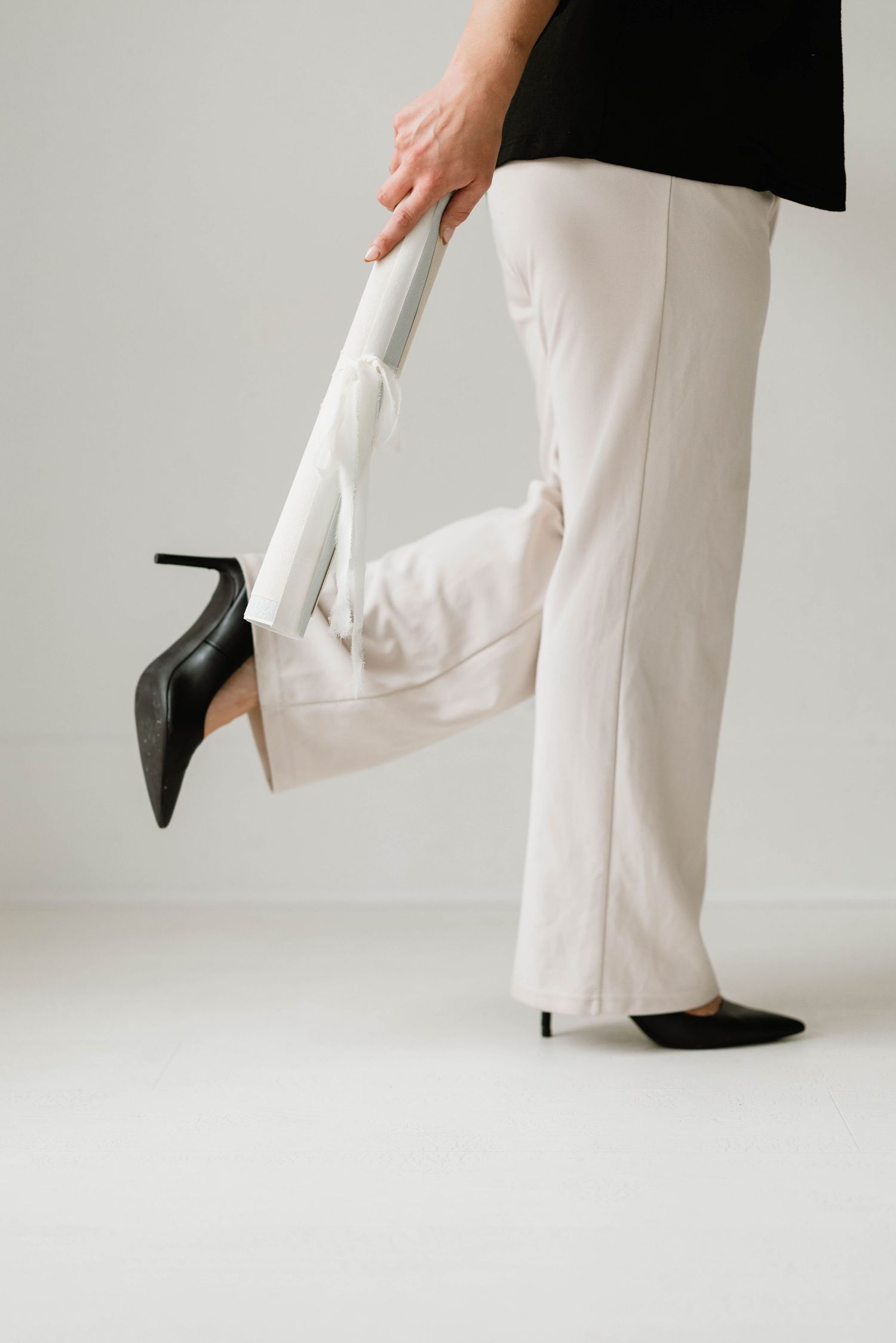 Woman in white wide-leg trousers and black heels lifting leg against light gray background.