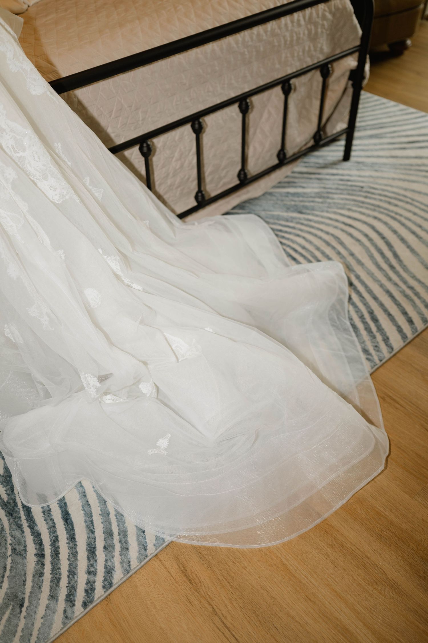 An elegant white wedding dress train draped over a striped rug beside a bed.