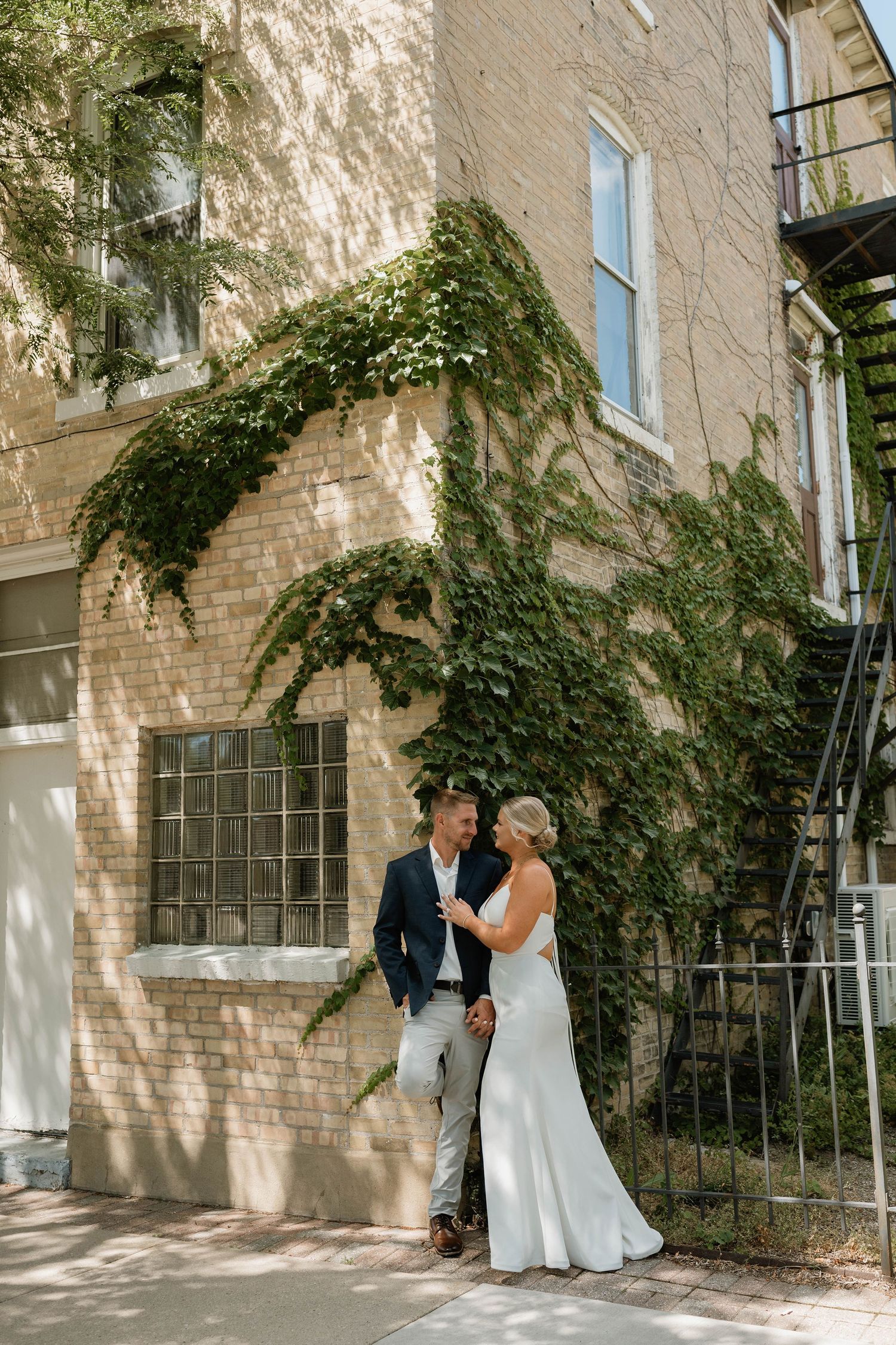 Bride and groom gazing at each other beside an ivy-covered brick building with iron staircase.
