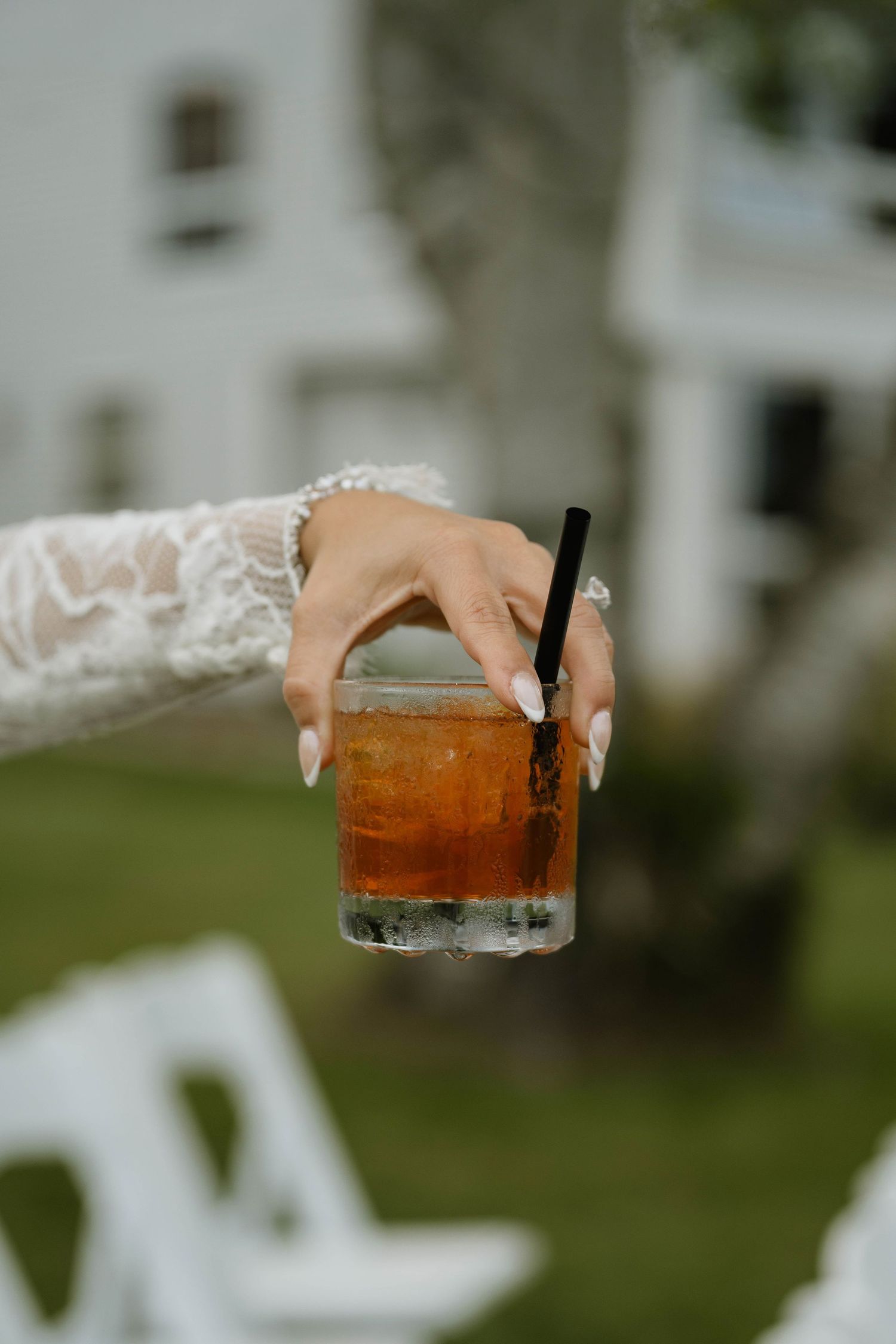 Bride in lace sleeve wedding dress holding iced cocktail with black straw outdoors.