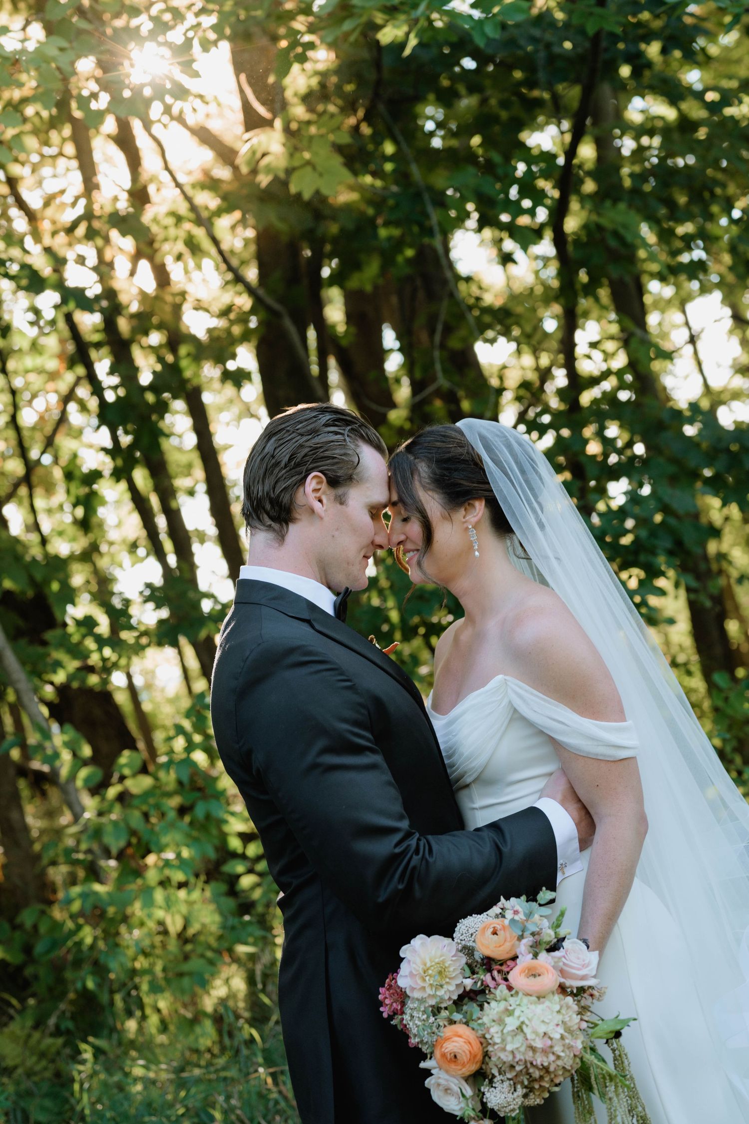 Bride and groom share a romantic forehead touch in a sunlit forest, bride holding a pastel bouquet in a white off-shoulder gown.
