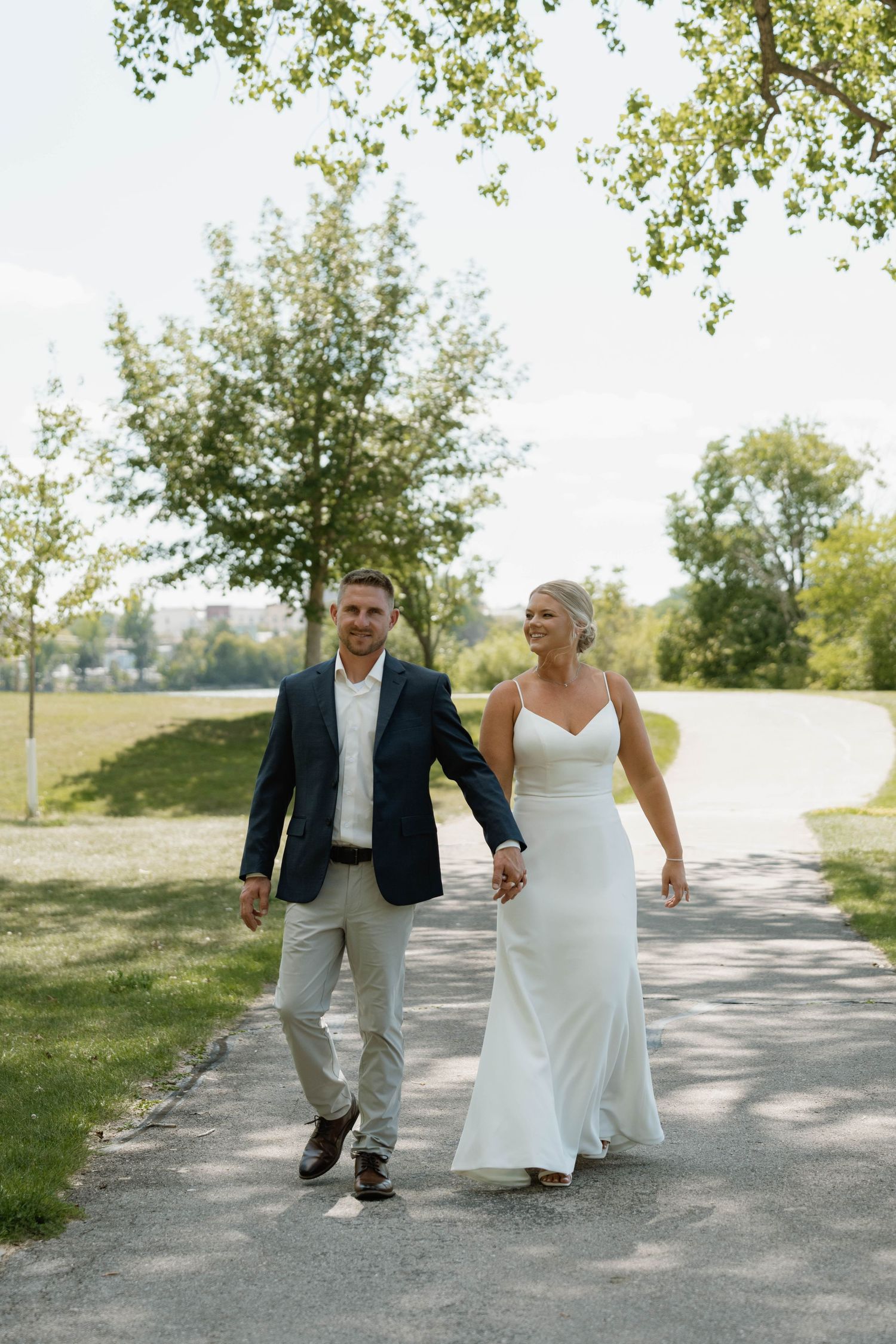 A bride and groom walk hand in hand along a sunny park path, she in a white gown, he in a navy blazer.